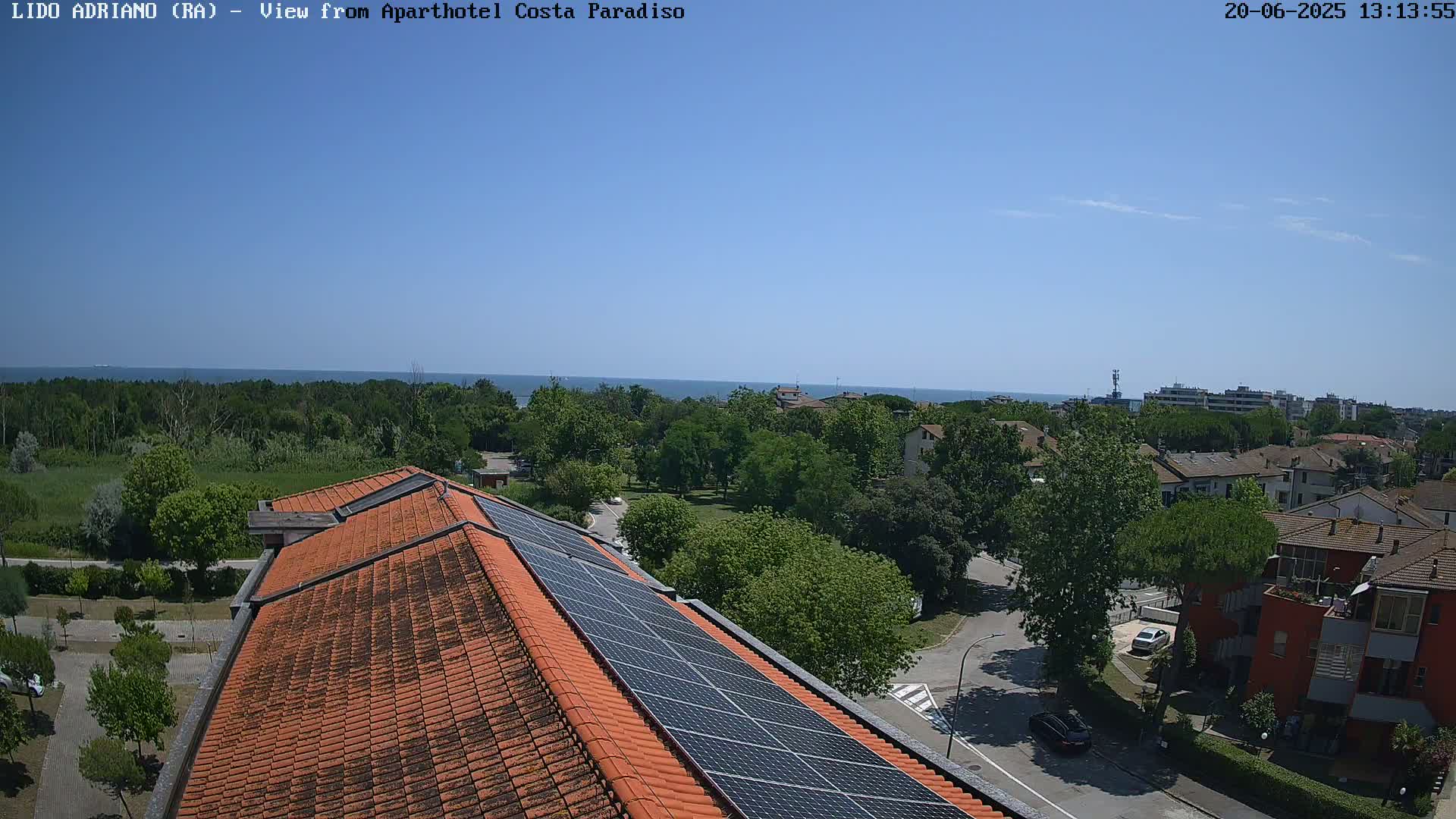 A rooftop with solar panels overlooks a sunny seaside town with a calm blue ocean in the distance.