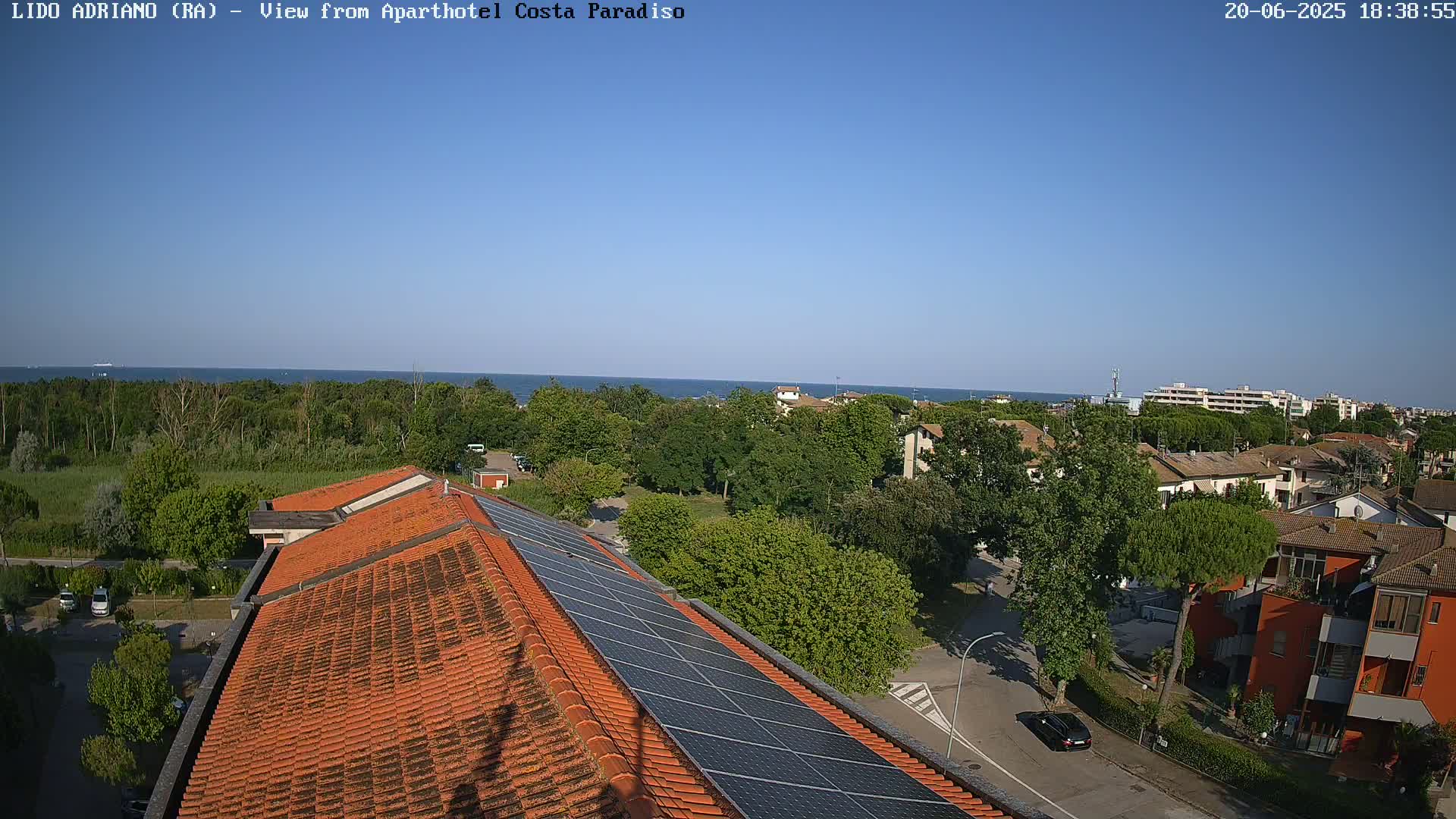 A rooftop with solar panels overlooks a residential area with green trees and a calm blue sea under a clear blue sky.