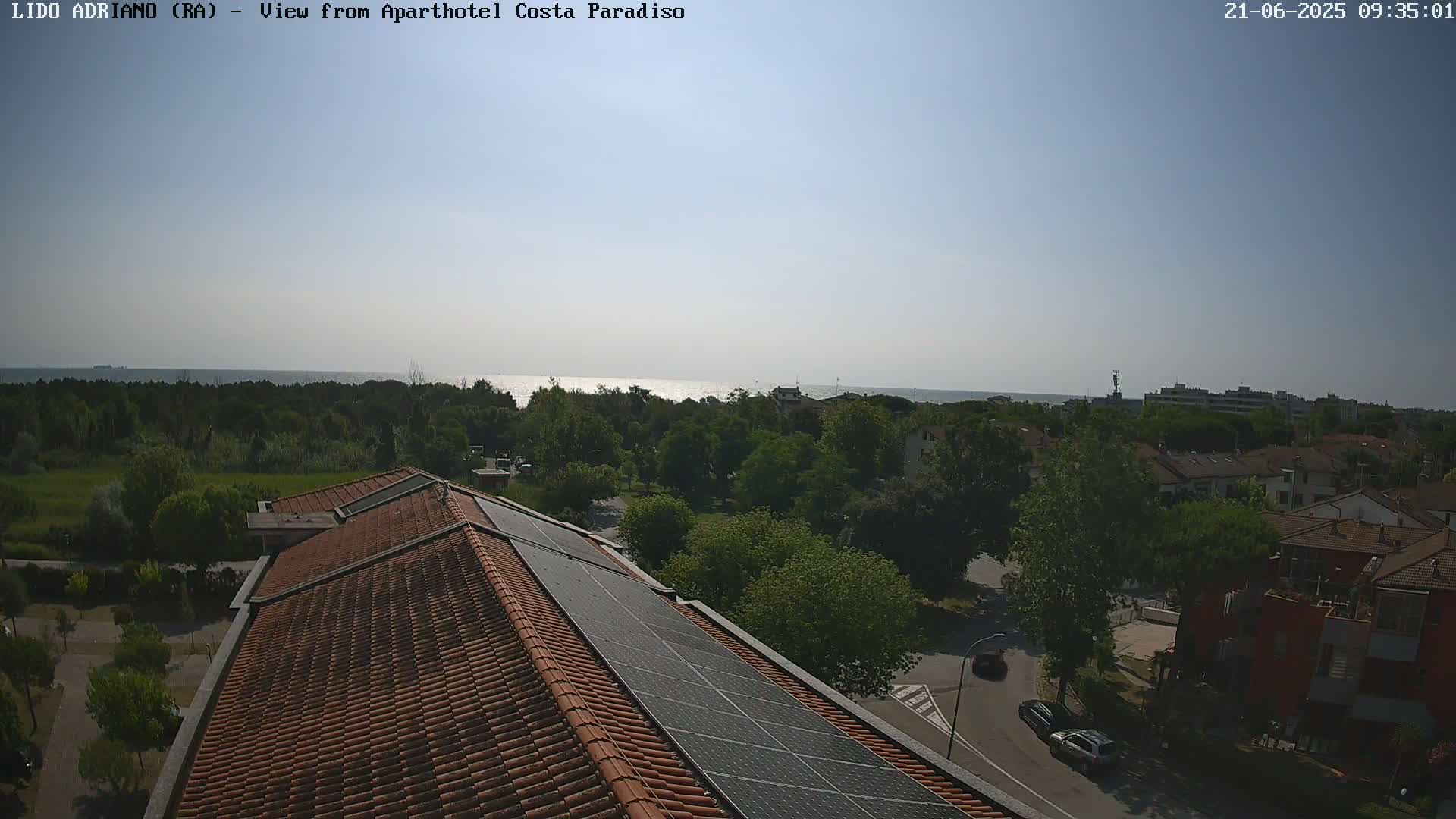 A mostly sunny view of a seaside town from a rooftop with solar panels, showing a calm sea in the distance.