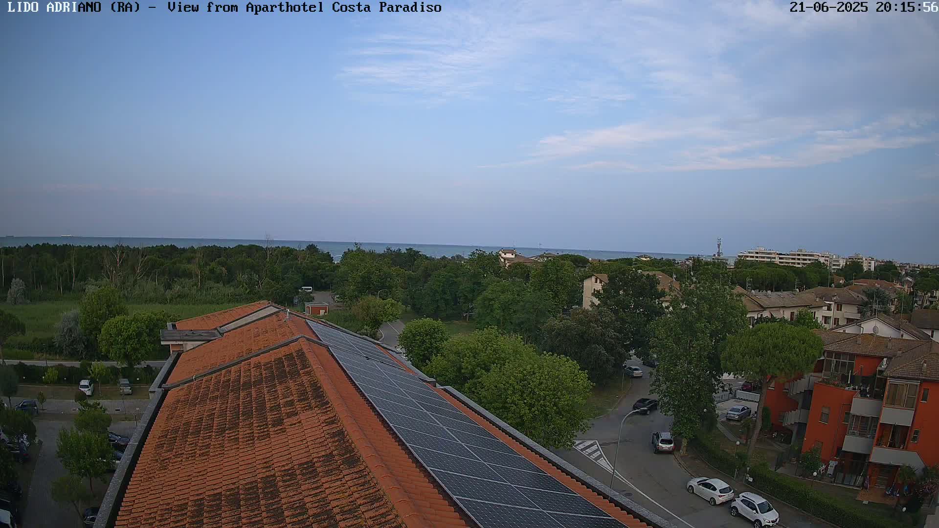 A mostly clear evening sky overlooks a coastal town with a red-tiled building featuring solar panels on its roof; cars are parked on the streets, and a calm sea is visible in the distance.