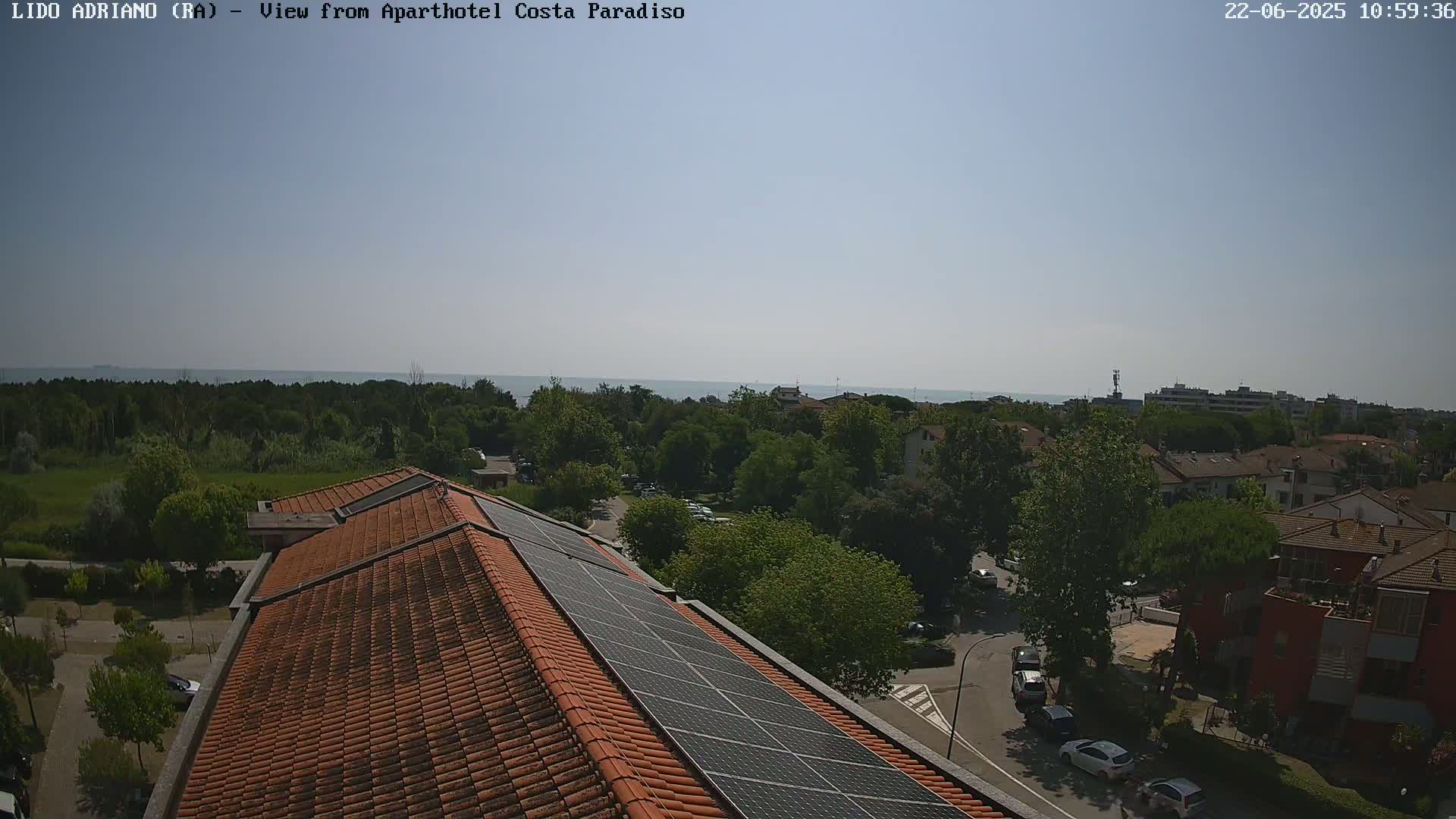 A clear, sunny day reveals a coastal residential area with a building rooftop featuring solar panels, overlooking a calm sea in the distance.