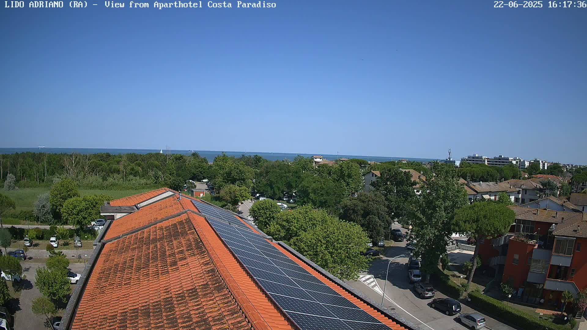 A rooftop view on a sunny day shows an orange tiled roof with solar panels, overlooking a residential area with lush green trees and a calm blue sea in the distance.
