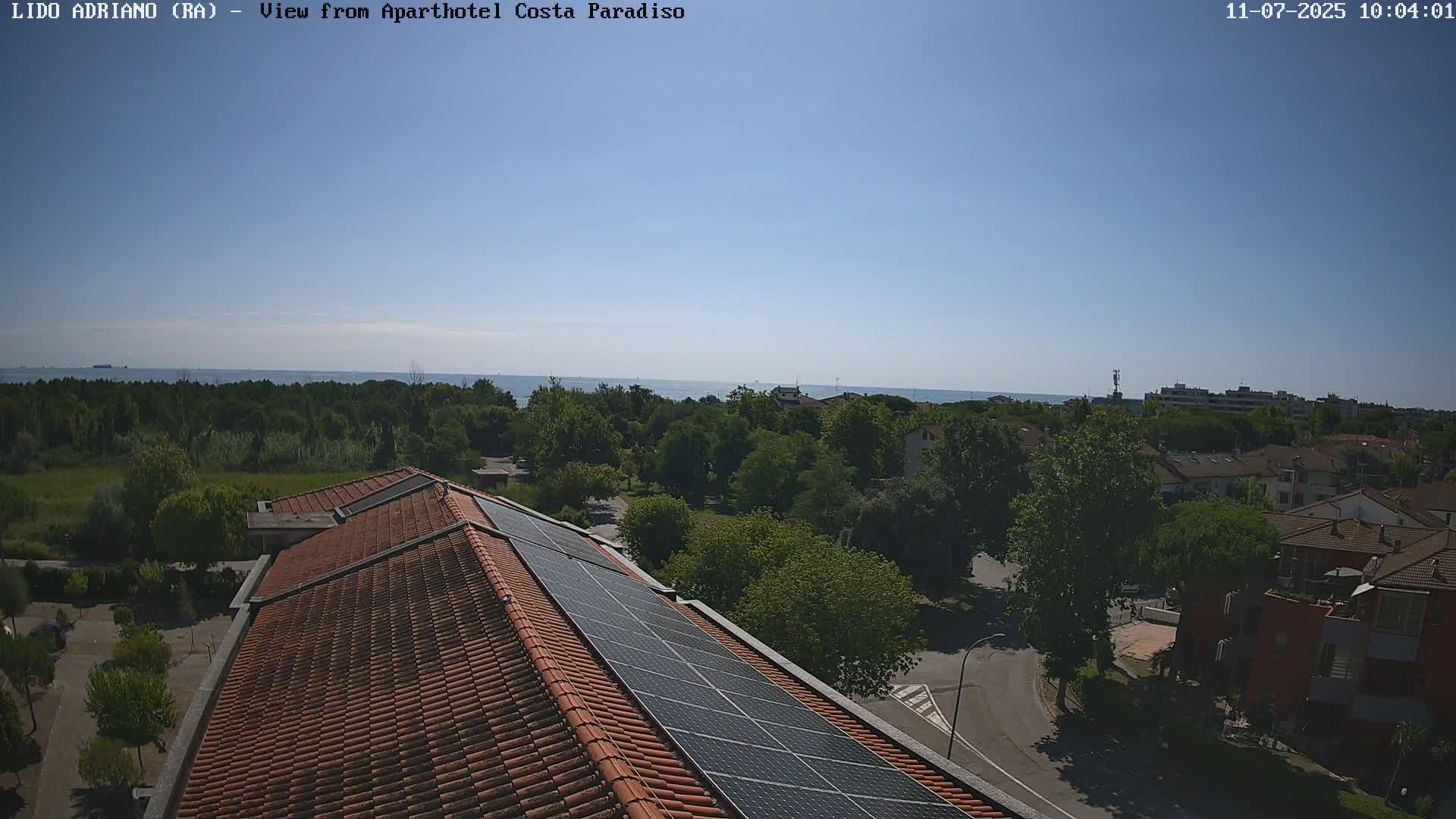 A rooftop with solar panels overlooks a residential area and a calm sea under a clear blue sky.