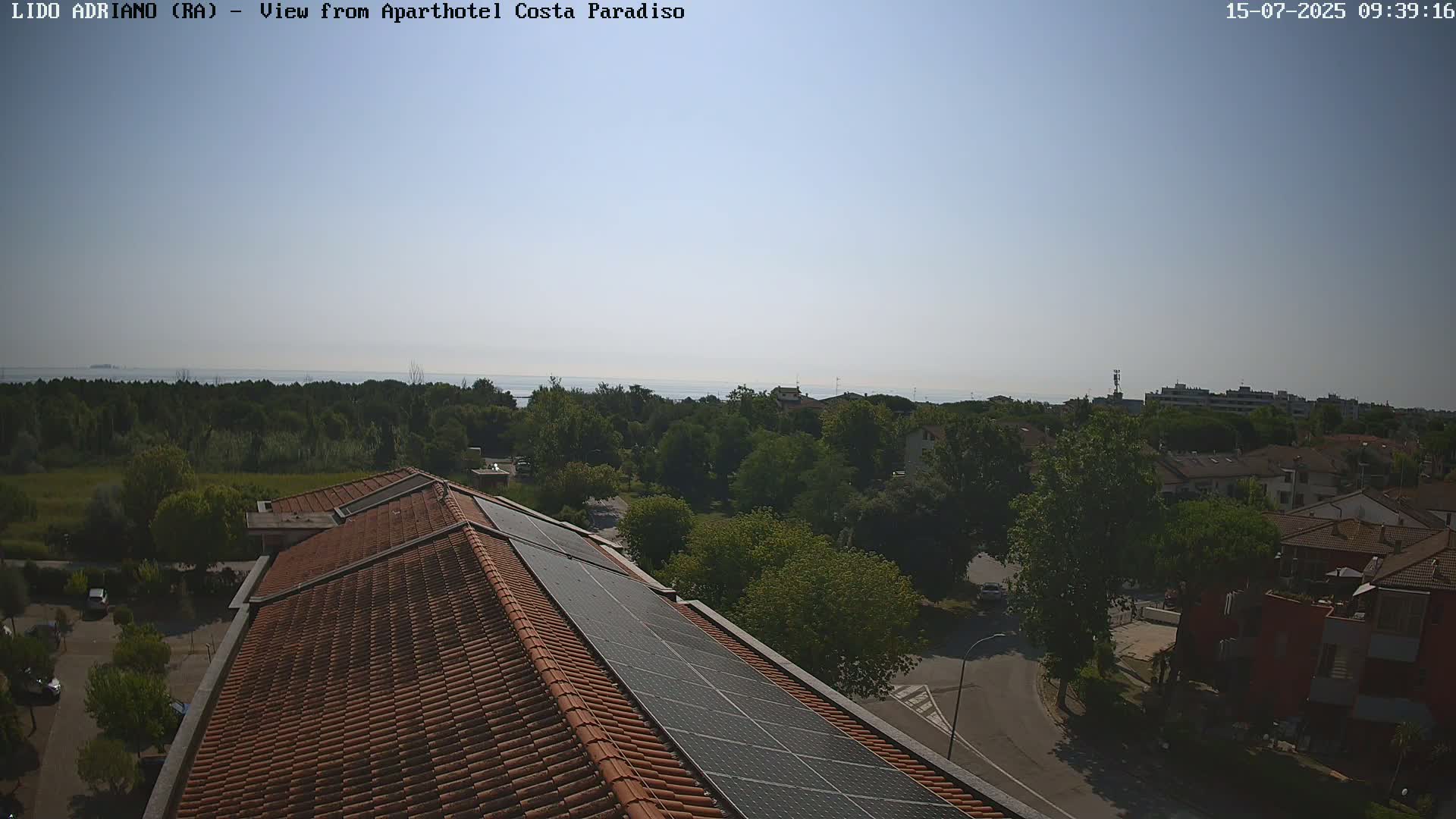 A rooftop with solar panels overlooks a tree-lined residential area and a calm sea under a clear sky.