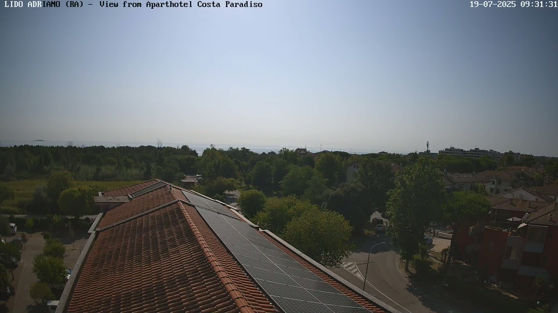 A rooftop with solar panels overlooks a tree-lined residential area and a calm sea under a clear sky.