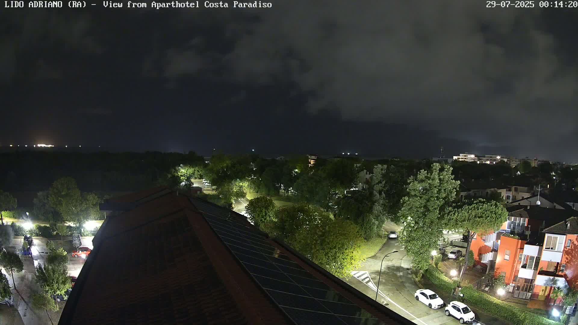 A nighttime view of a residential area with overcast skies shows a few cars on wet roads, and trees and buildings illuminated by streetlights.