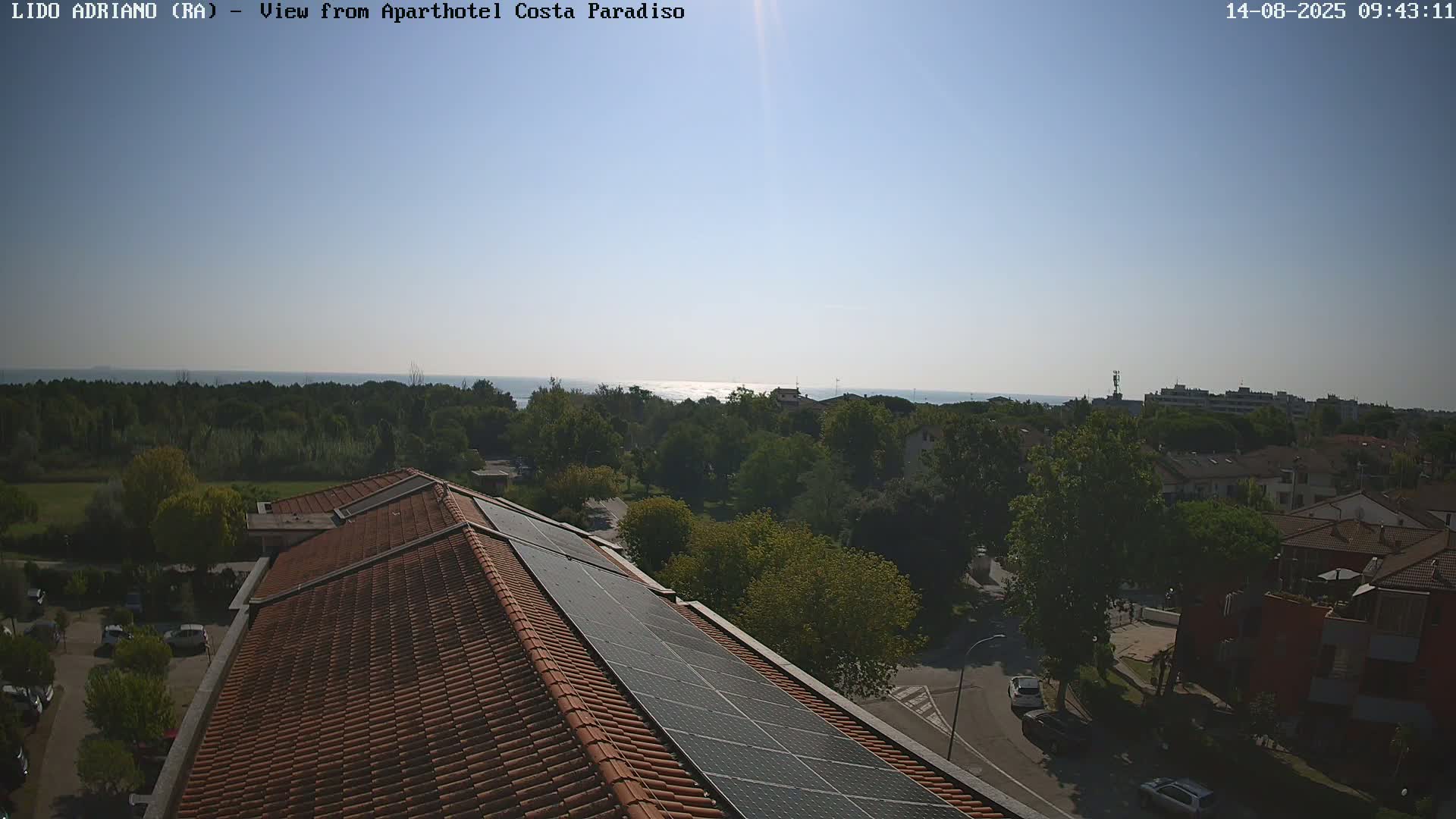 A sunny view from a rooftop with solar panels shows a calm sea in the distance, trees, buildings, and a road below.