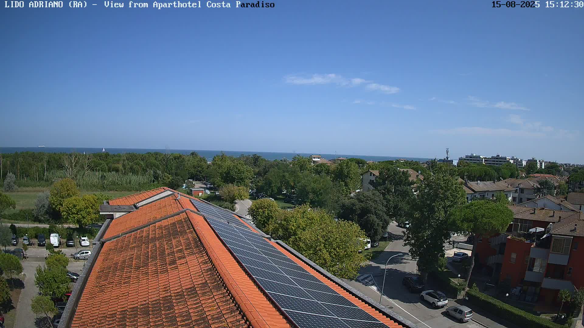 A mostly sunny day overlooking a tiled roof with solar panels, a tree-lined area, and a calm body of water in the distance.