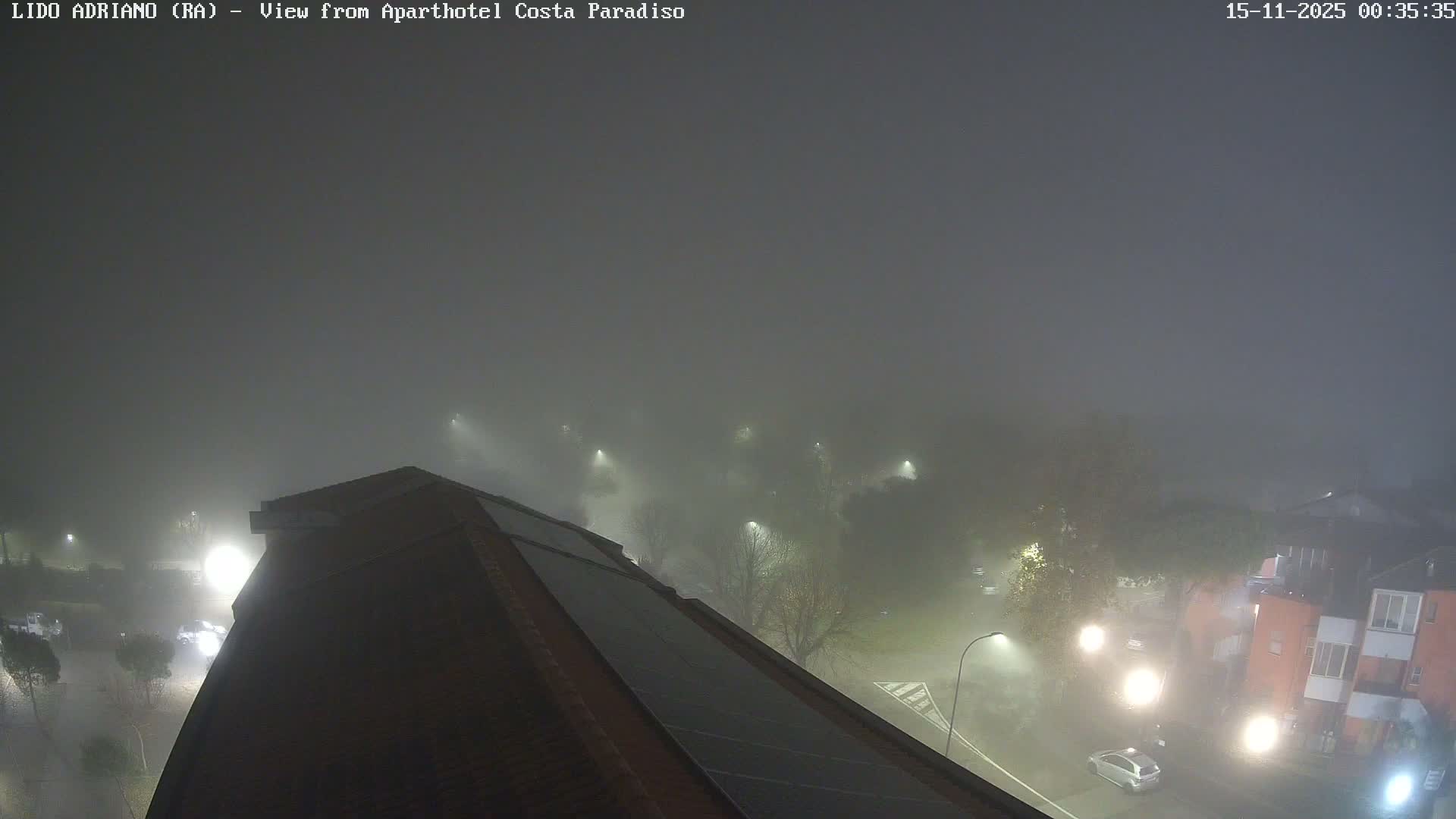 A dense nighttime fog obscures an elevated view of a neighborhood, revealing a roof with solar panels in the foreground, glowing streetlights illuminating hazy streets with cars, and indistinct trees and buildings.