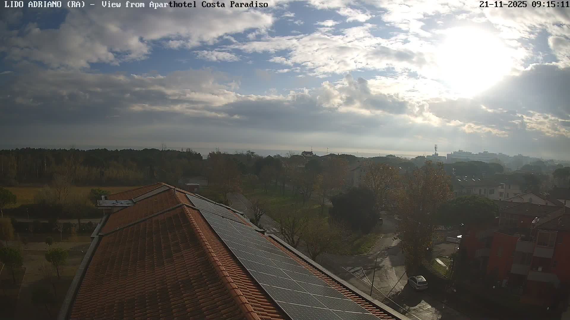 The image shows a sunny, partly cloudy daytime view over a coastal town featuring a foreground rooftop with solar panels, a mix of residential buildings and abundant trees, and a distant sea horizon.