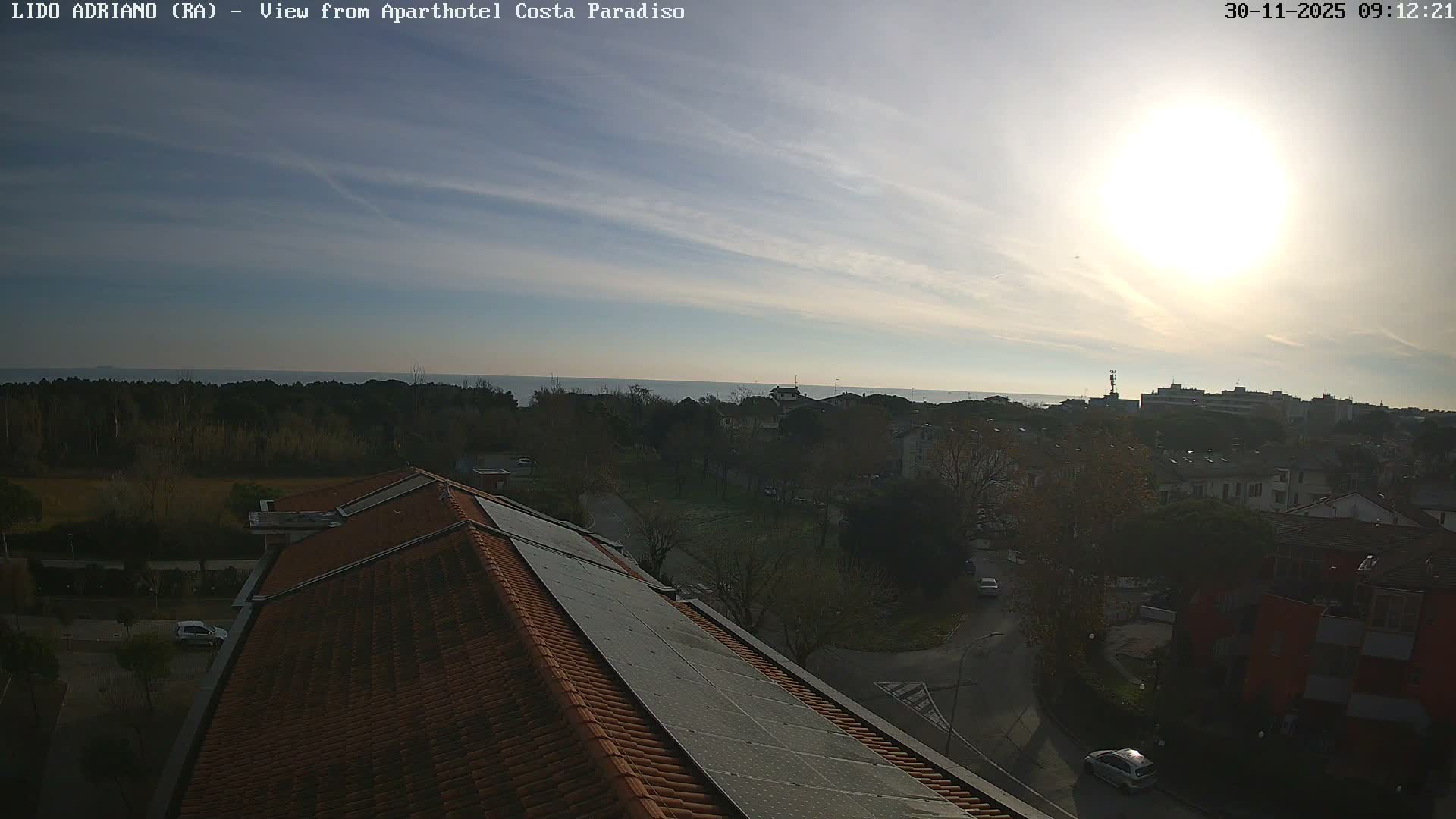 The image captures a bright and sunny day over a coastal town, with a tiled rooftop adorned with solar panels in the foreground, leading to a cityscape, a treeline, and the calm sea under a partly cloudy blue sky.