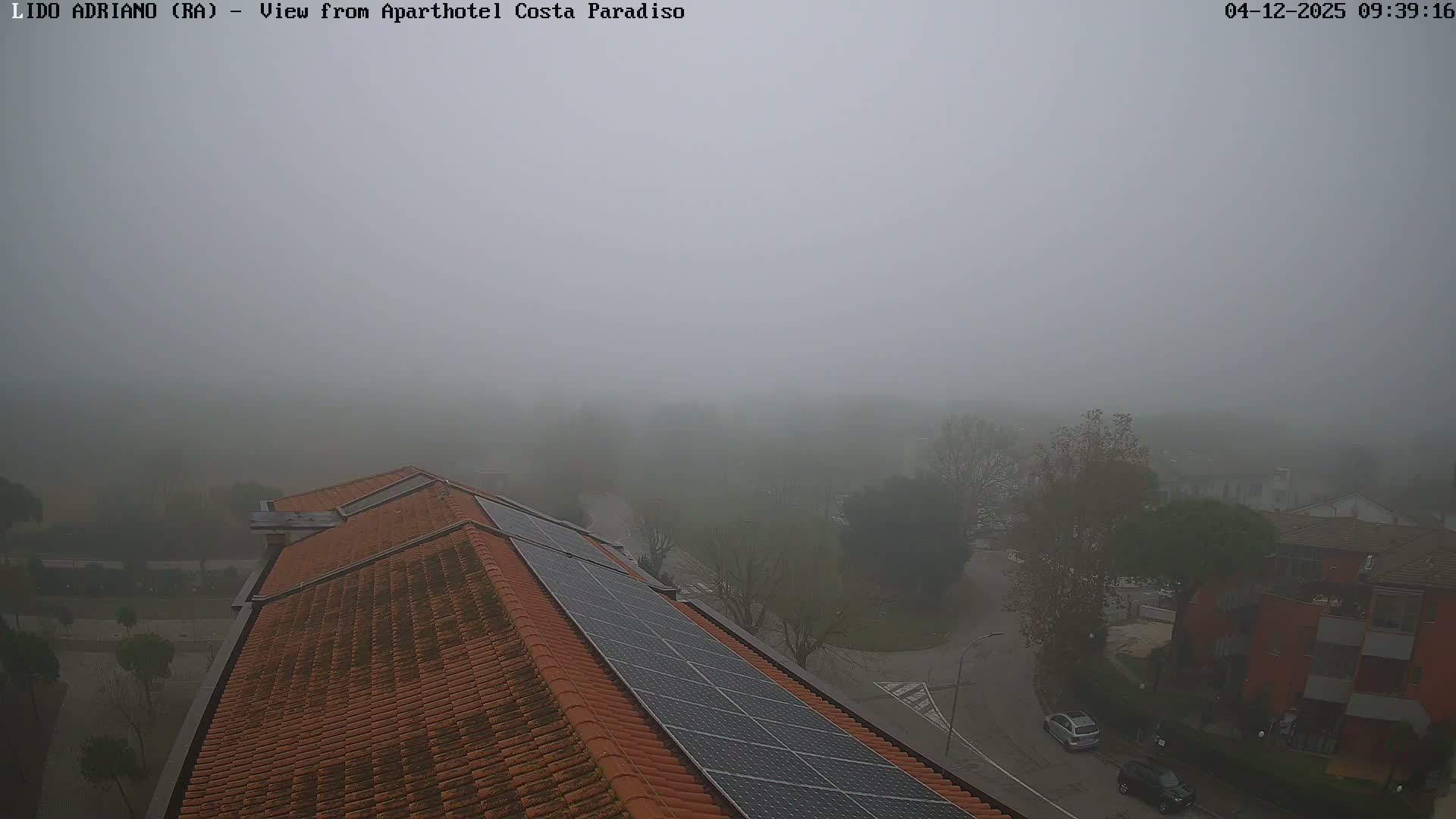 A dense fog envelops an outdoor scene from a high vantage point, revealing solar panels on a terracotta-tiled roof in the foreground, with a street, cars, and buildings dimly visible through the heavy mist.