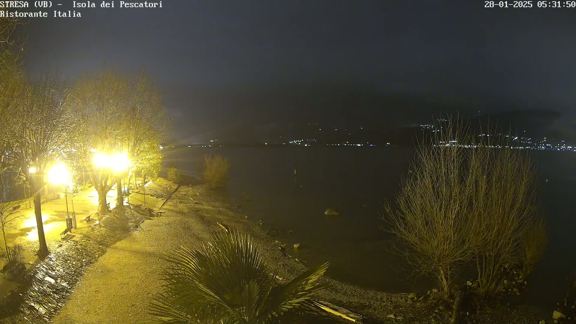 A nighttime lakeside scene shows a wet, pebble shoreline with sparsely leafed trees and illuminated walkways under a dark, possibly overcast sky.