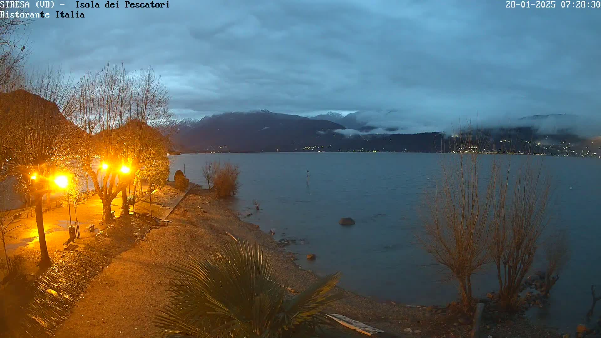 A shoreline at night, lit by streetlights, overlooks a calm lake with distant mountains shrouded in fog under a cloudy sky.