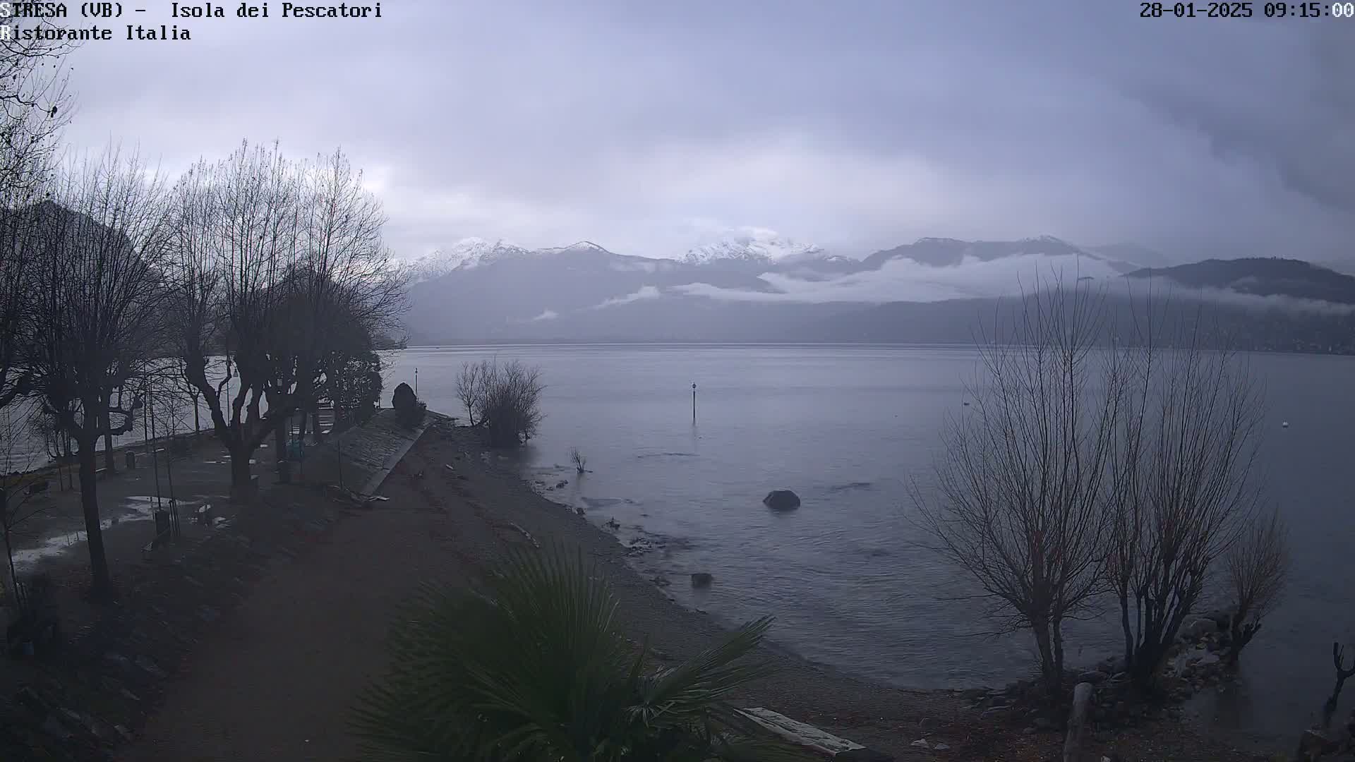 A grey, overcast day shows a calm lake with snow-capped mountains in the background, bare trees lining the shoreline, and a pebble beach in the foreground.