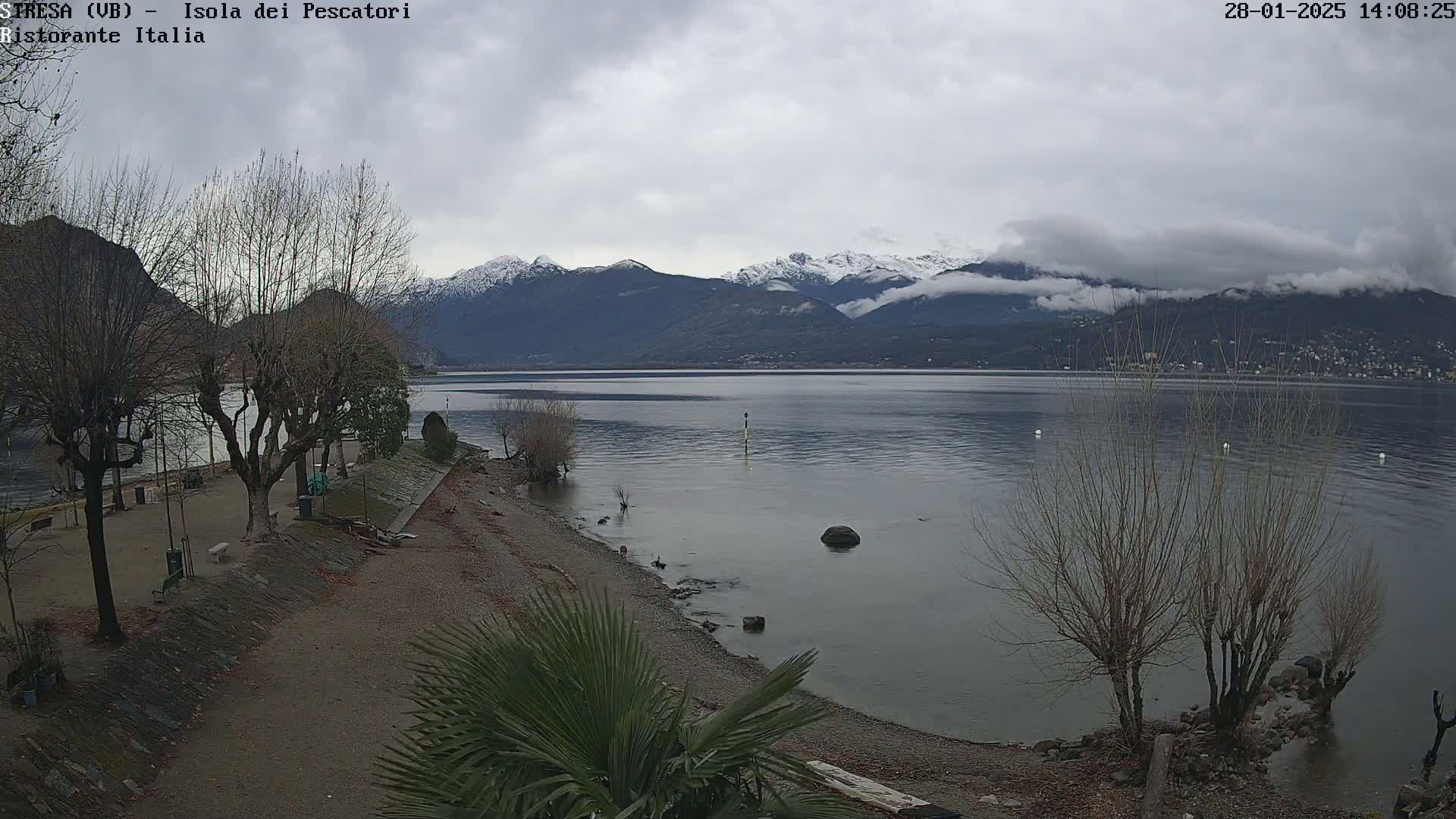 A mostly cloudy day shows a calm lake with snow-capped mountains in the background, a pebble beach in the foreground, and bare trees along the shoreline.