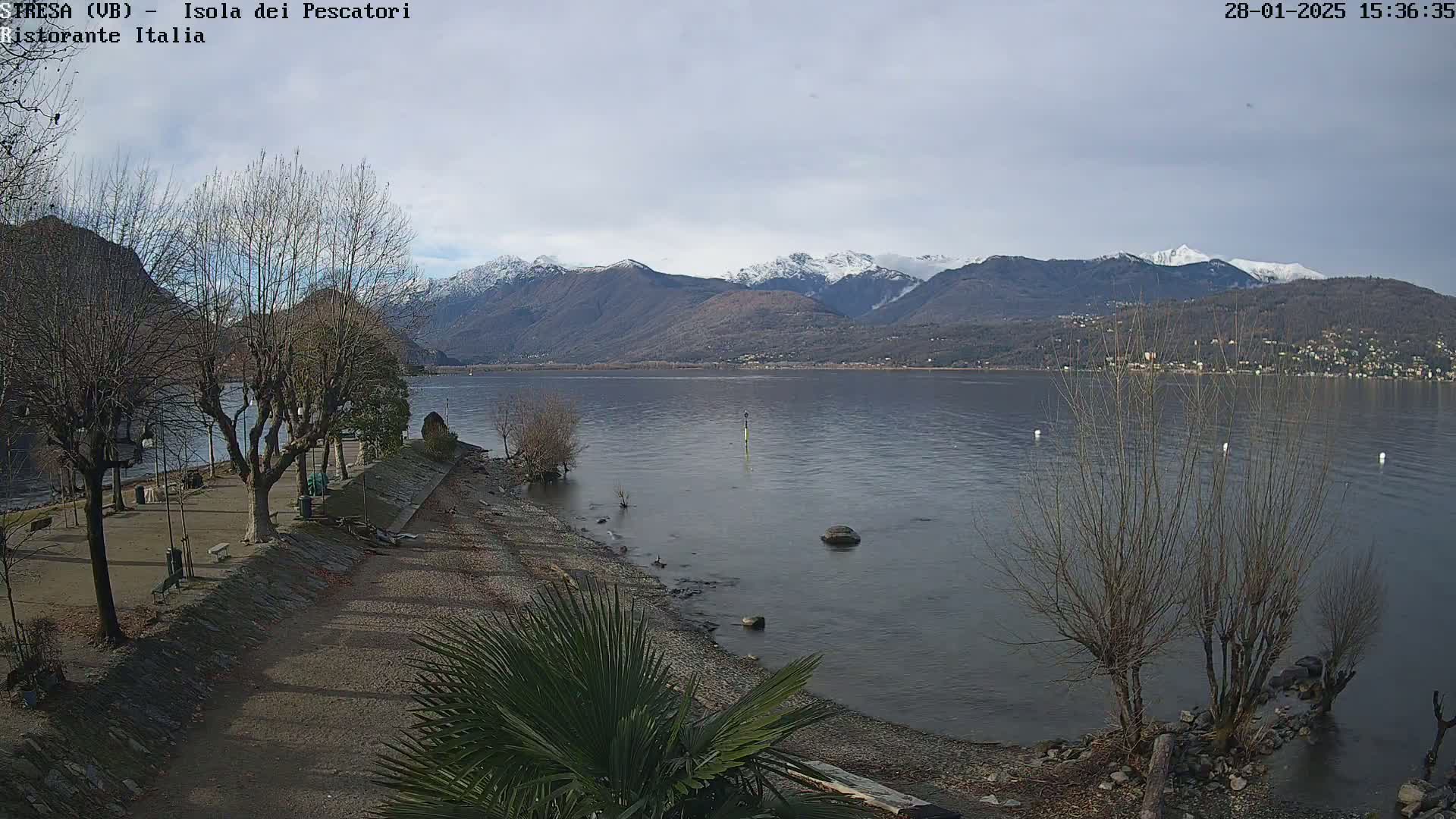 A calm lake stretches out to snow-capped mountains under a mostly cloudy sky, with bare trees lining a shoreline path.
