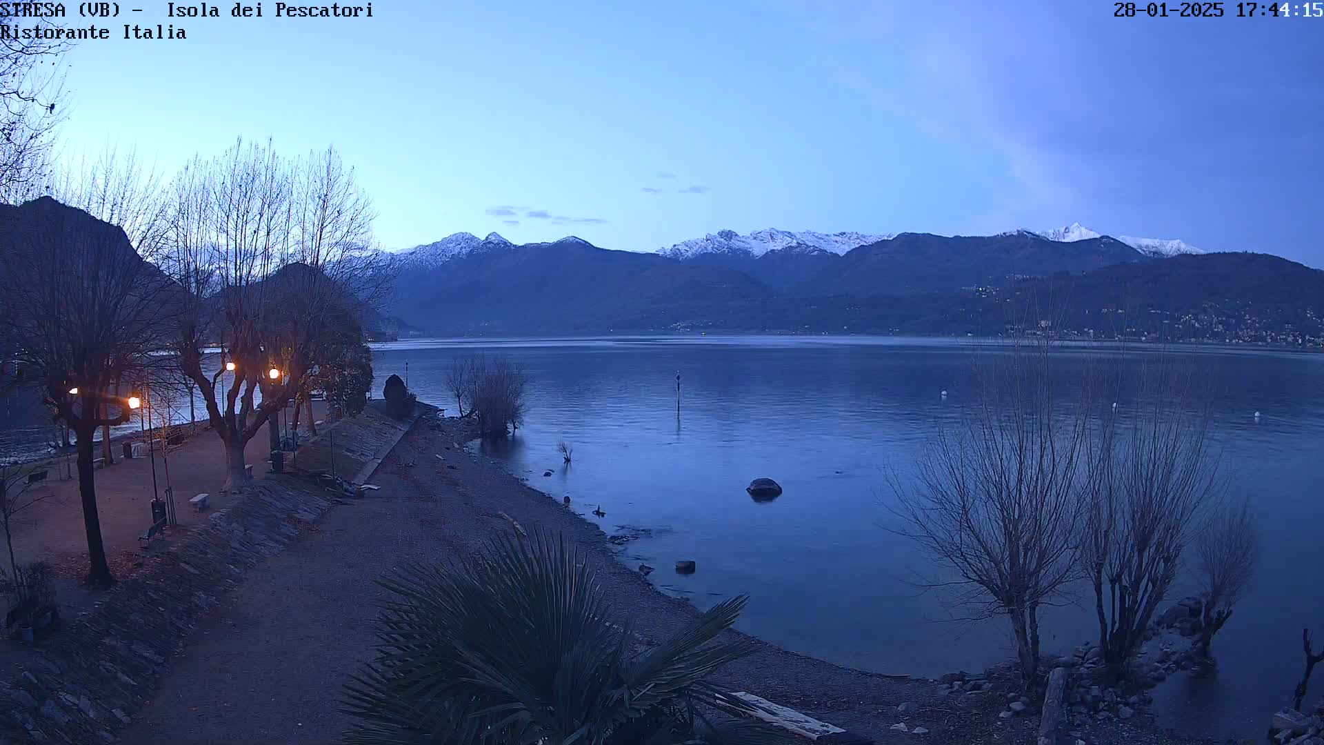A twilight view of a calm lake, snow-capped mountains in the background, and a shoreline with bare trees and lit lampposts.