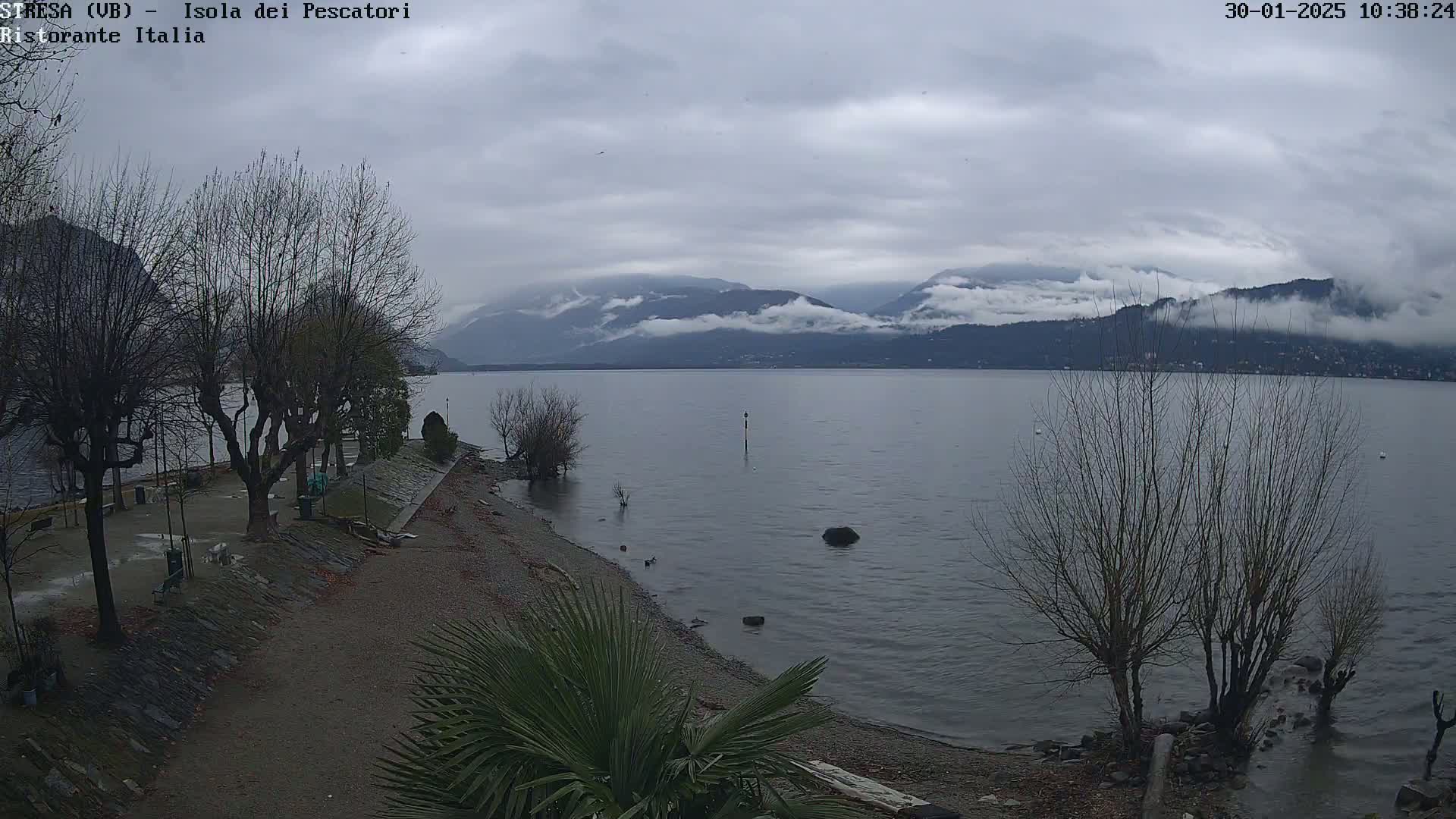 A view of a calm lake with leafless trees lining a pebble shoreline, under a cloudy sky and mountains in the distance.