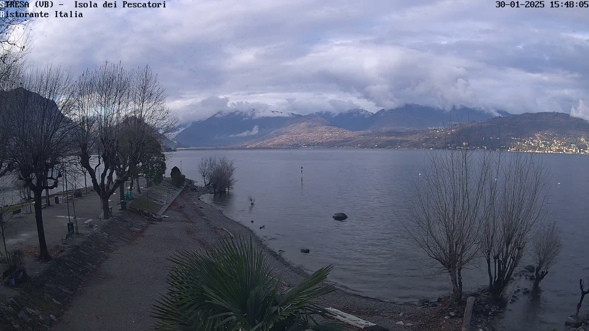 A partly cloudy day shows a shoreline with bare trees and a calm lake extending to distant mountains.