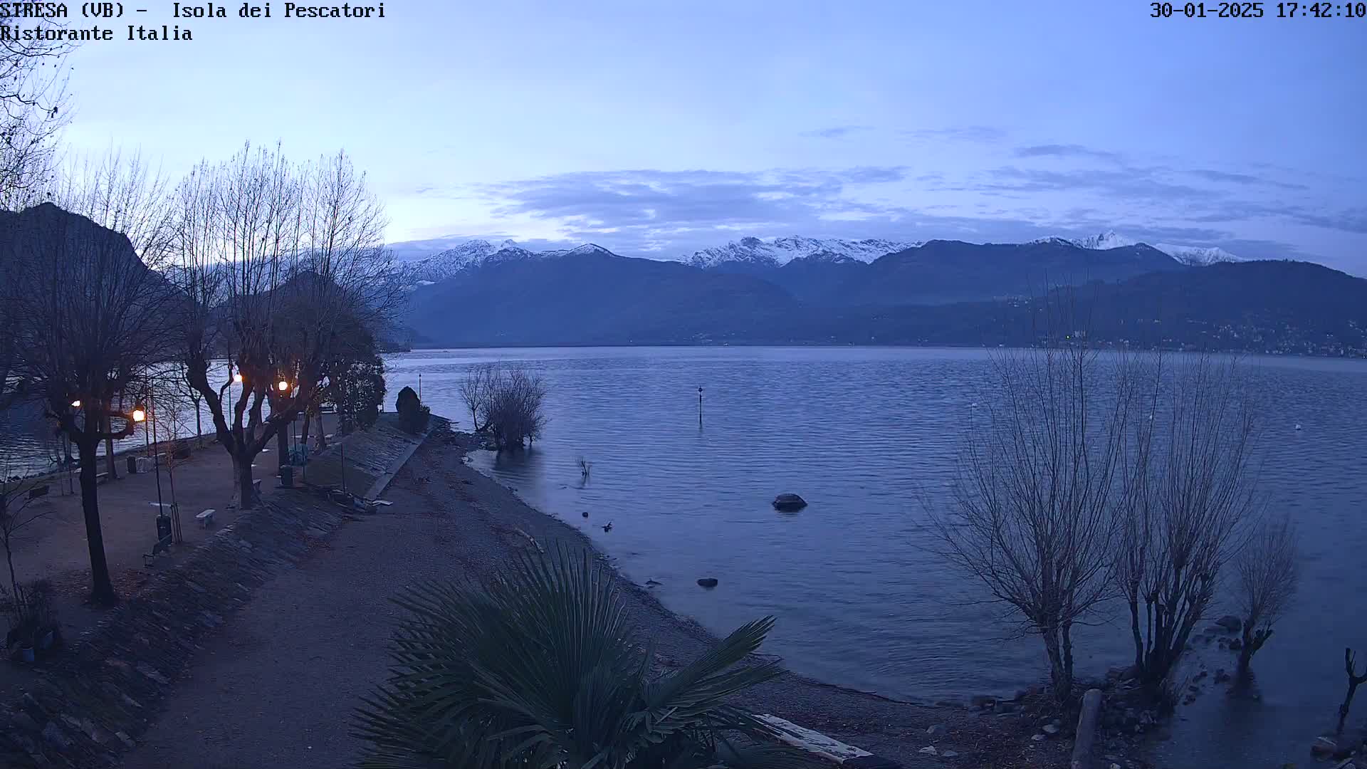A calm lake at dusk, under a twilight sky, is bordered by a paved waterfront path lined with bare trees and lit by streetlights, with snow-capped mountains visible in the distance.