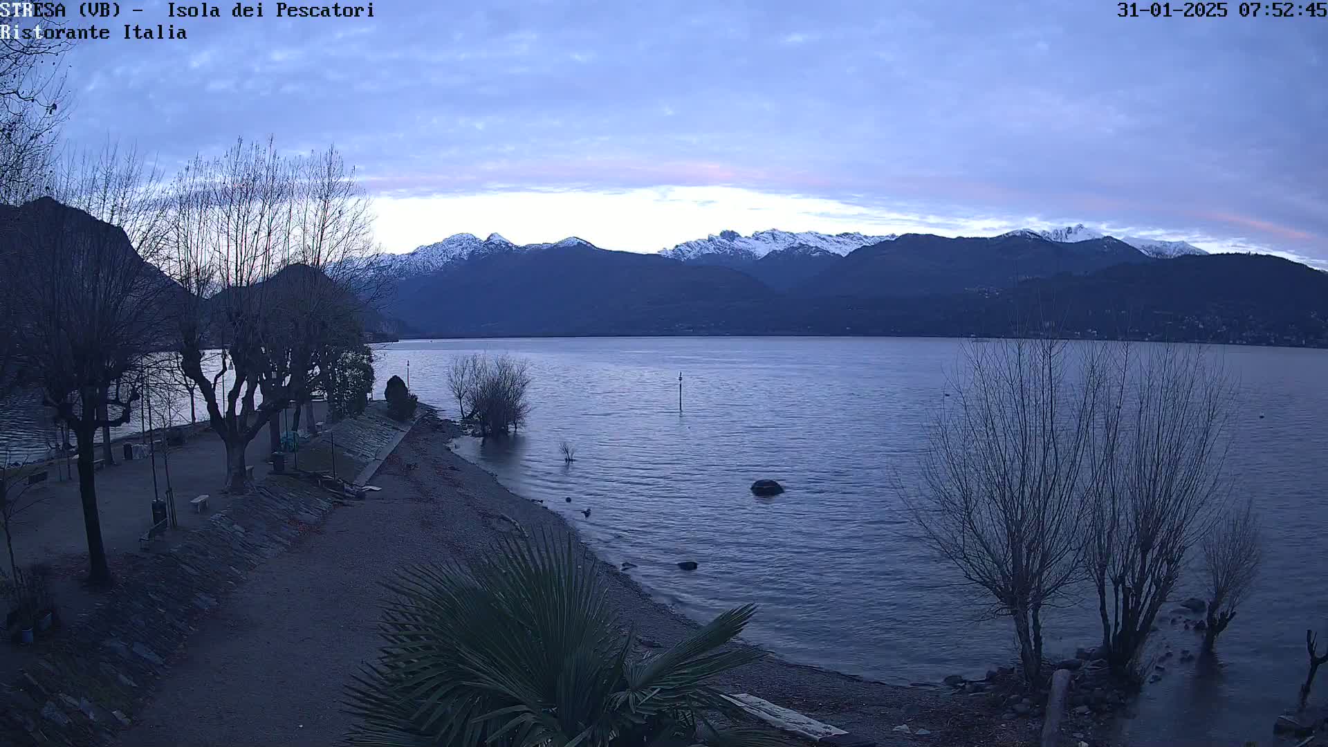 A calm lake at dawn is backed by snow-capped mountains under a mostly clear sky.