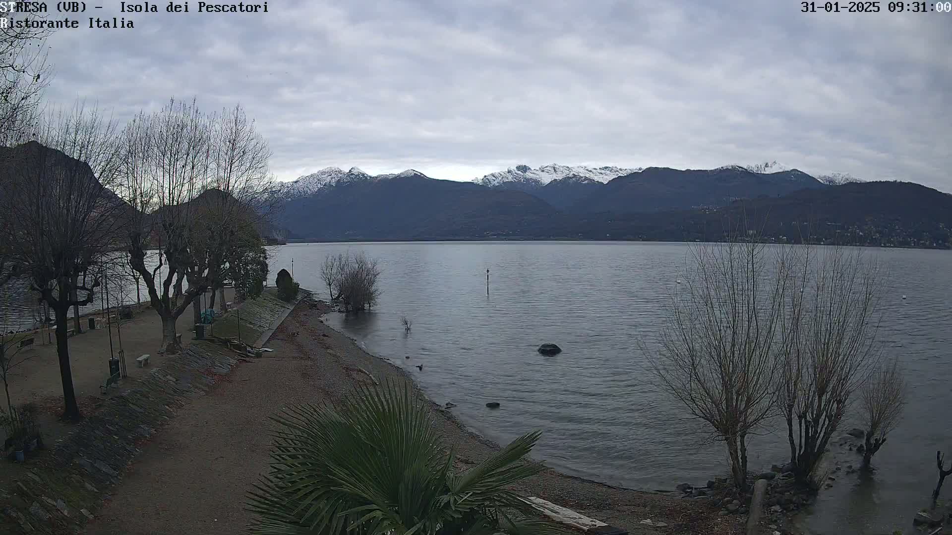 A calm lake stretches towards snow-capped mountains under a cloudy sky, with leafless trees lining a pebbled shoreline.