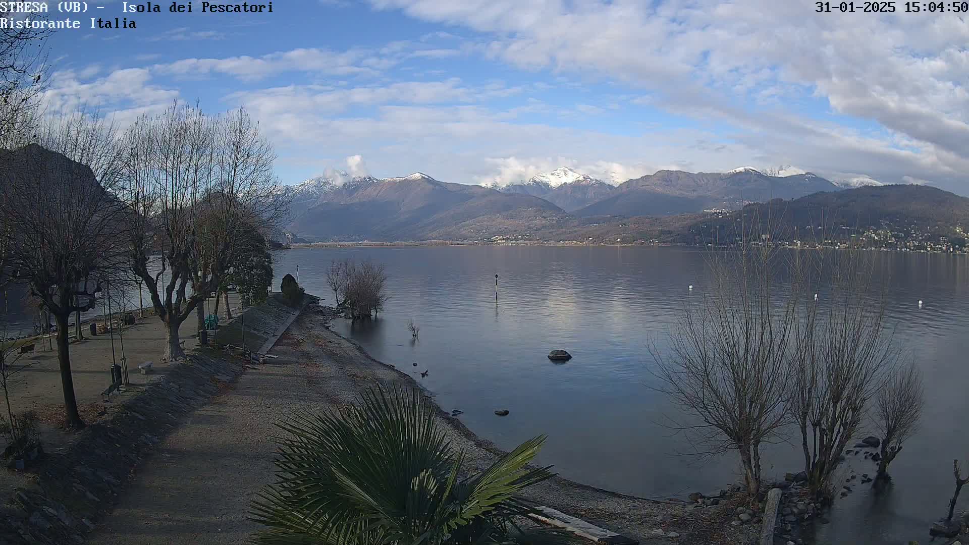A partly cloudy day reveals a tranquil lake scene, with snow-capped mountains in the background and leafless trees lining a shoreline with a gravel beach.