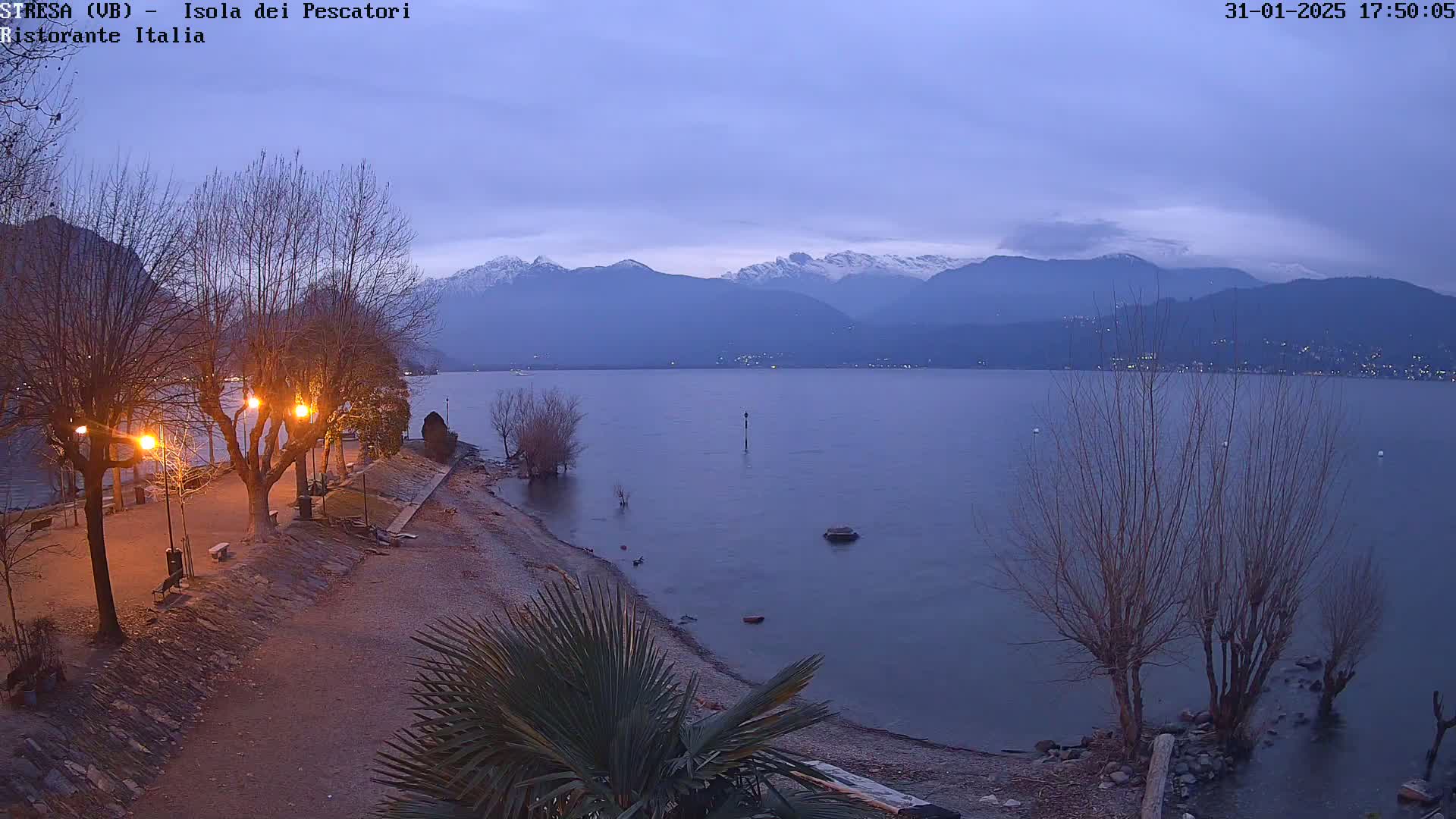 A calm lake at dusk, under a mostly clear sky, is backed by snow-capped mountains and a shoreline with bare trees and lights.