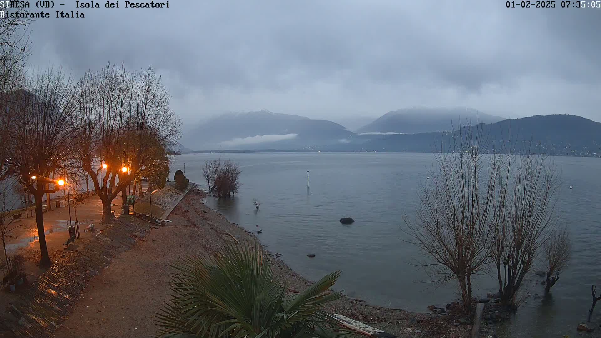 A calm lake under an overcast sky, seen from a shoreline with bare trees and lit lampposts, mountains in the distance.