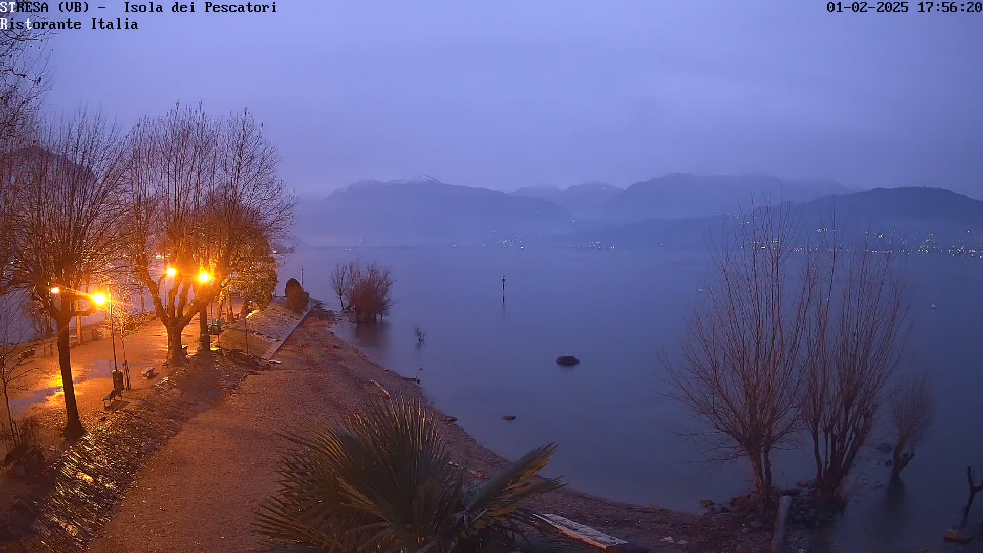A twilight view shows a lakeshore with bare trees, lit lampposts, and a calm lake extending to distant mountains under a dusky sky.