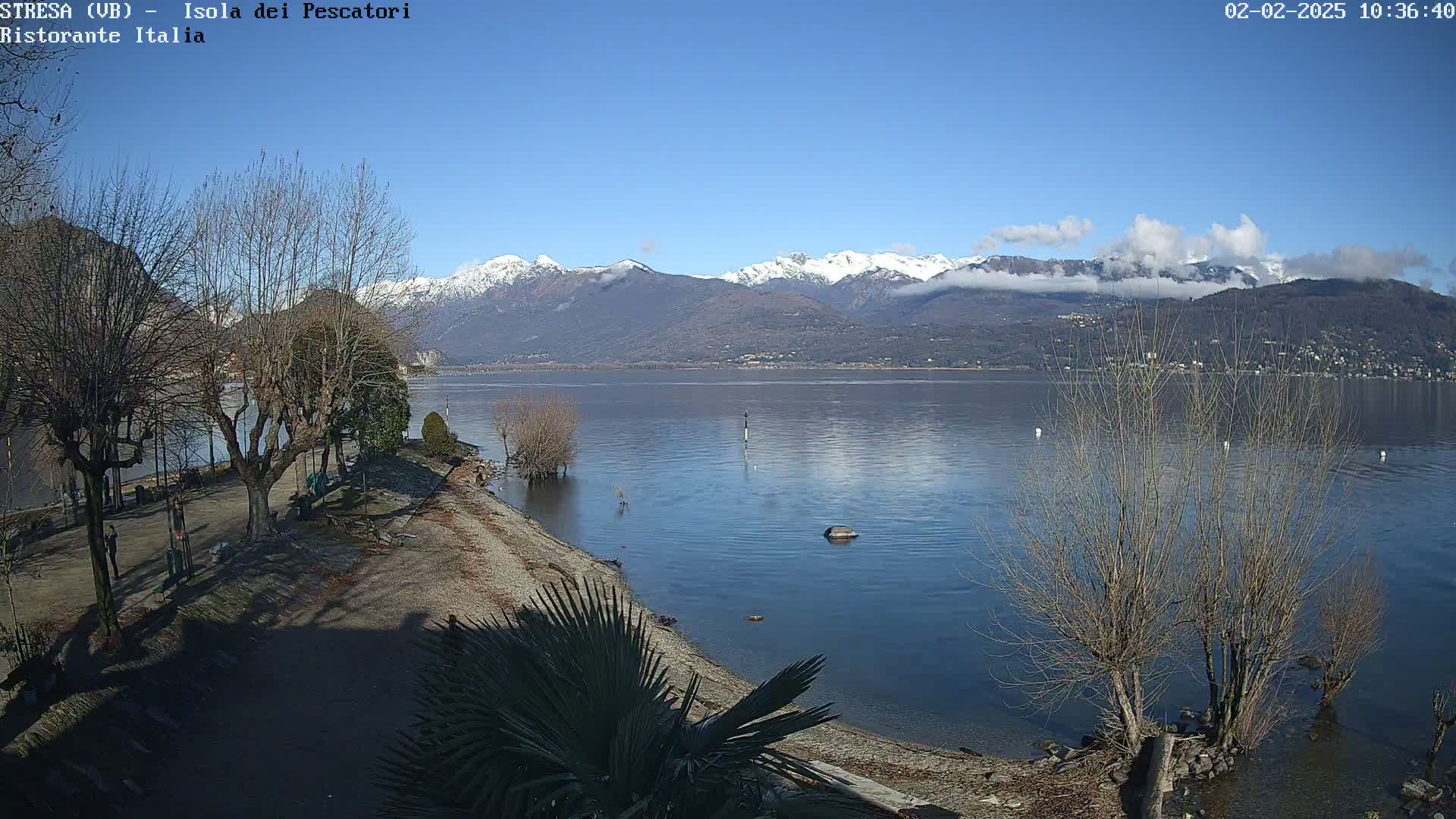 A partly cloudy sunny day reveals a lake shore with leafless trees, a sandy beach, and snow-capped mountains in the distance.