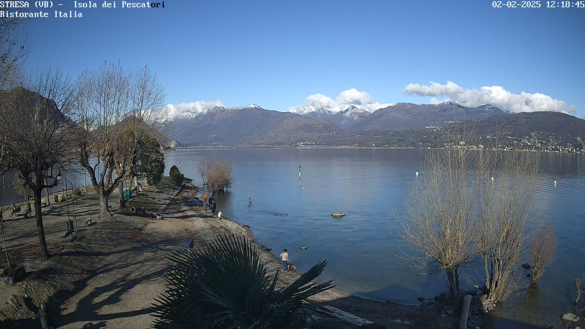 A sunny day shows snow-capped mountains across a calm lake, with bare trees lining a shoreline where a few people stroll.