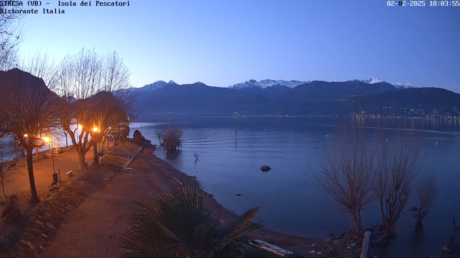 A twilight view of a lakeshore with bare trees, lit pathways, and snow-capped mountains in the distance.