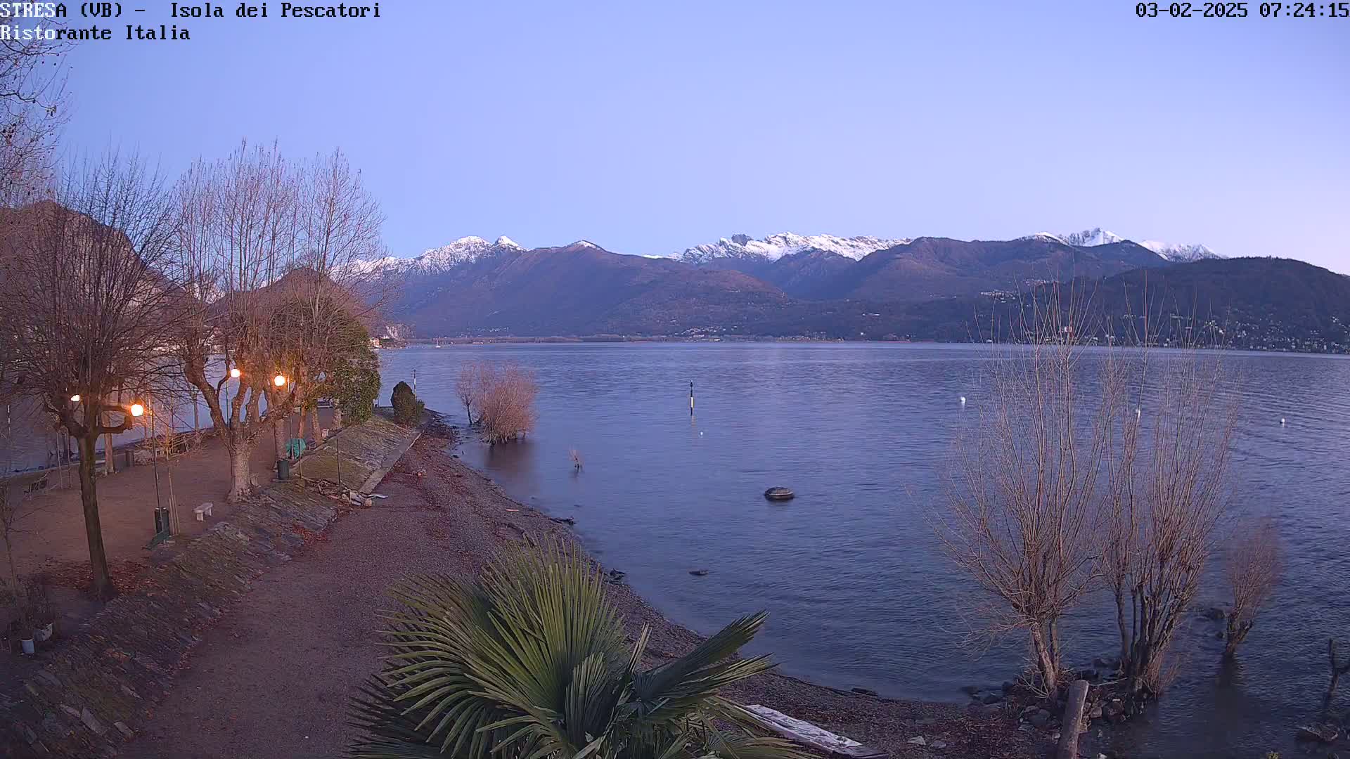 A calm lake at dawn is seen from a lakeside park with leafless trees and some lights, and snow-capped mountains are in the background.