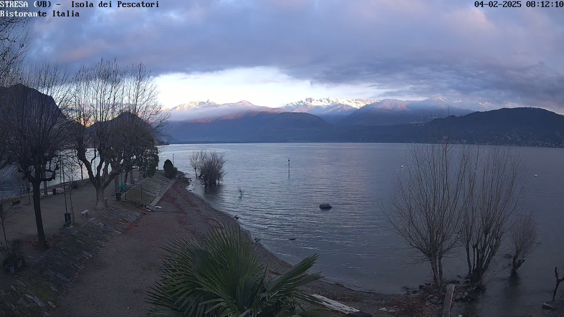 A calm lake with snow-capped mountains in the background under a cloudy sky, viewed from a lakeside park with bare trees.