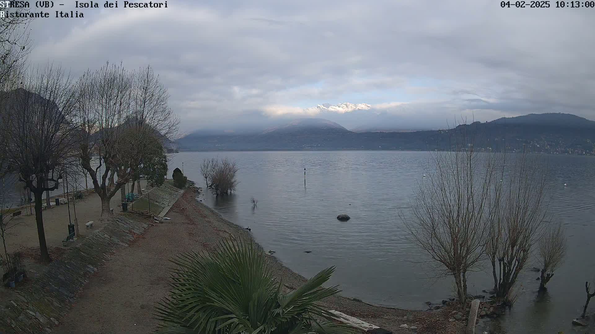 A mostly cloudy day reveals a lake shore with leafless trees and a calm lake, mountains in the background partially obscured by clouds.