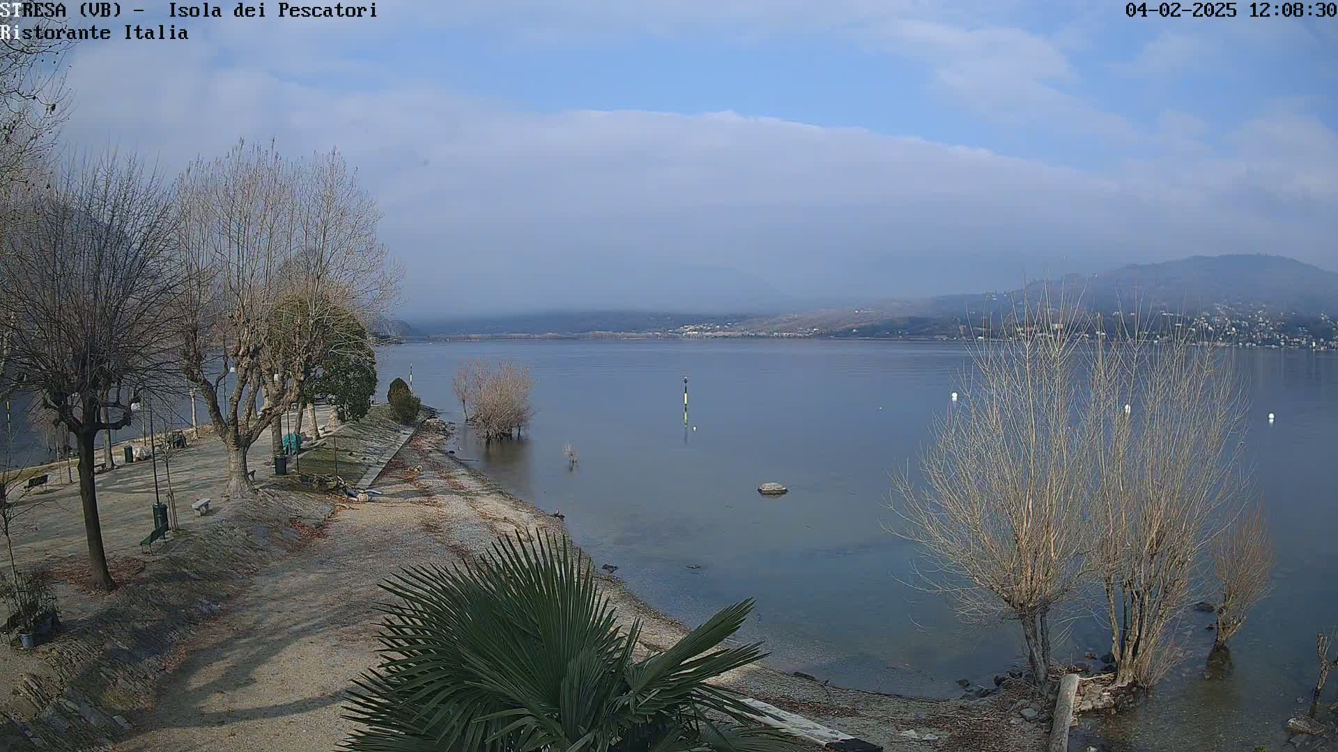A mostly sunny day reveals a calm lake with a shoreline featuring bare trees, a paved walkway, and a few benches, all under a partly cloudy sky.