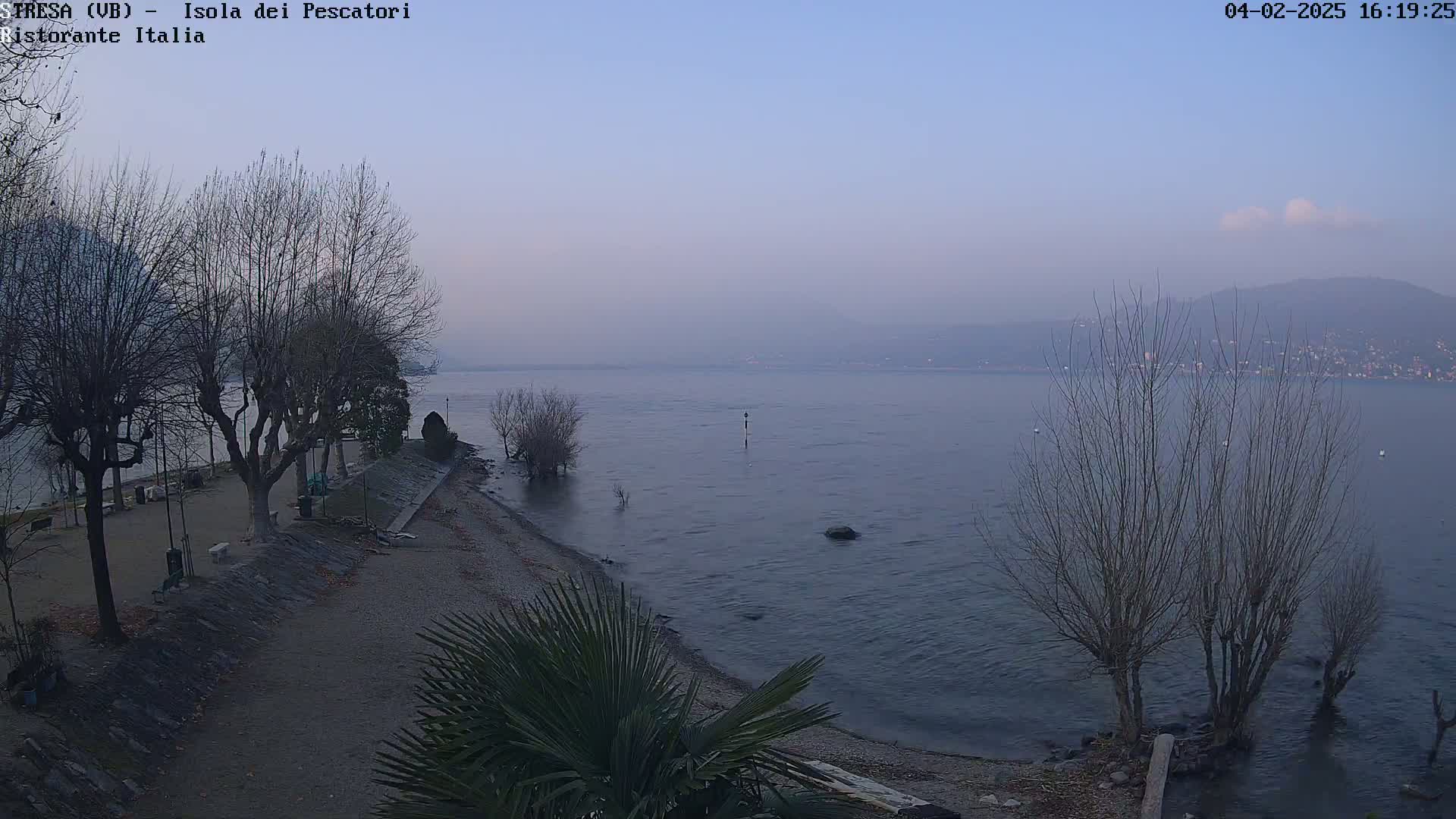 A hazy, dusky view of a calm lake stretches to distant mountains, with leafless trees lining a shoreline next to a gravel path.