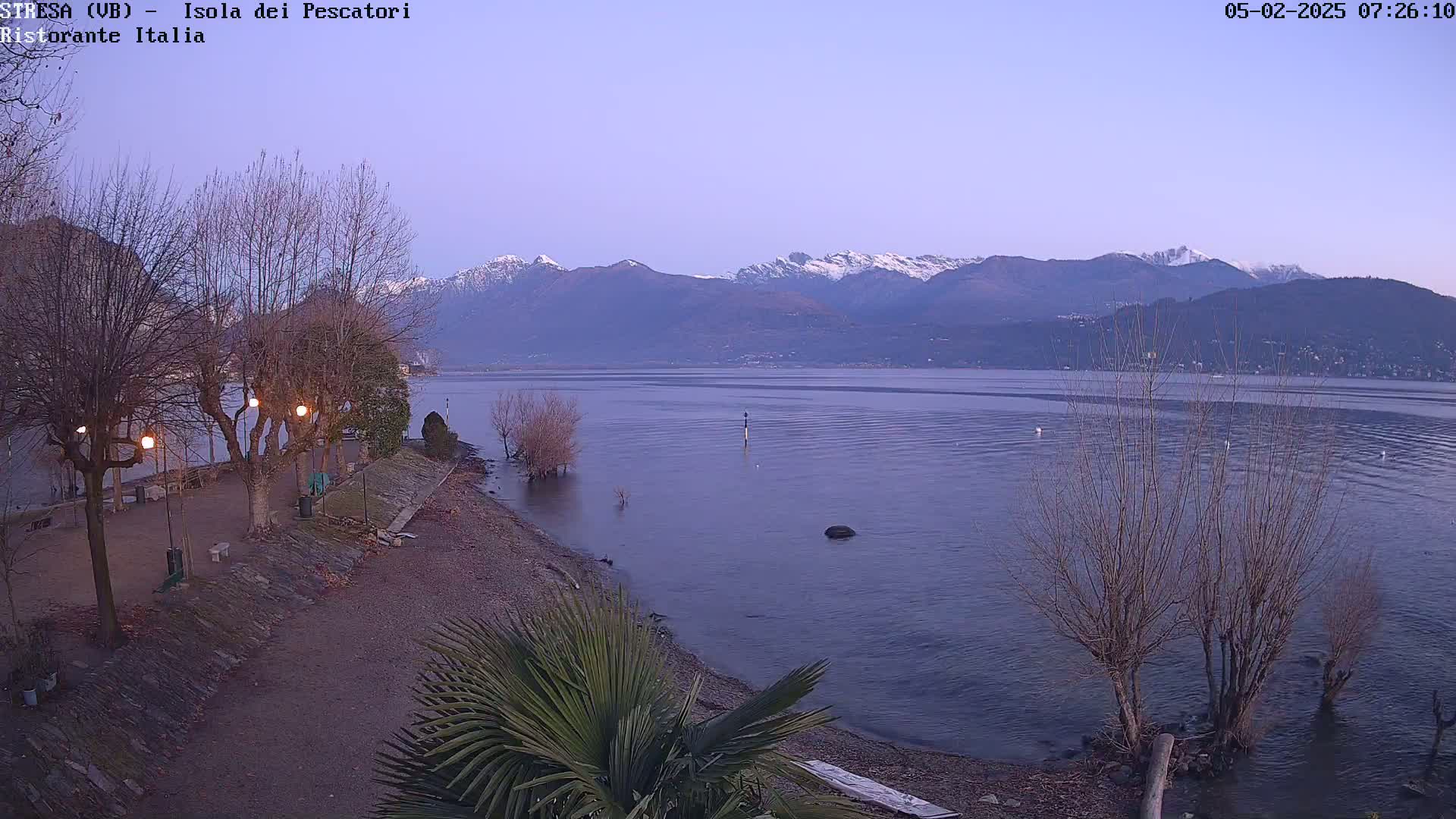 A calm lake at dawn, with snow-capped mountains in the background, is viewed from a lakeside park with bare trees and a few lights.