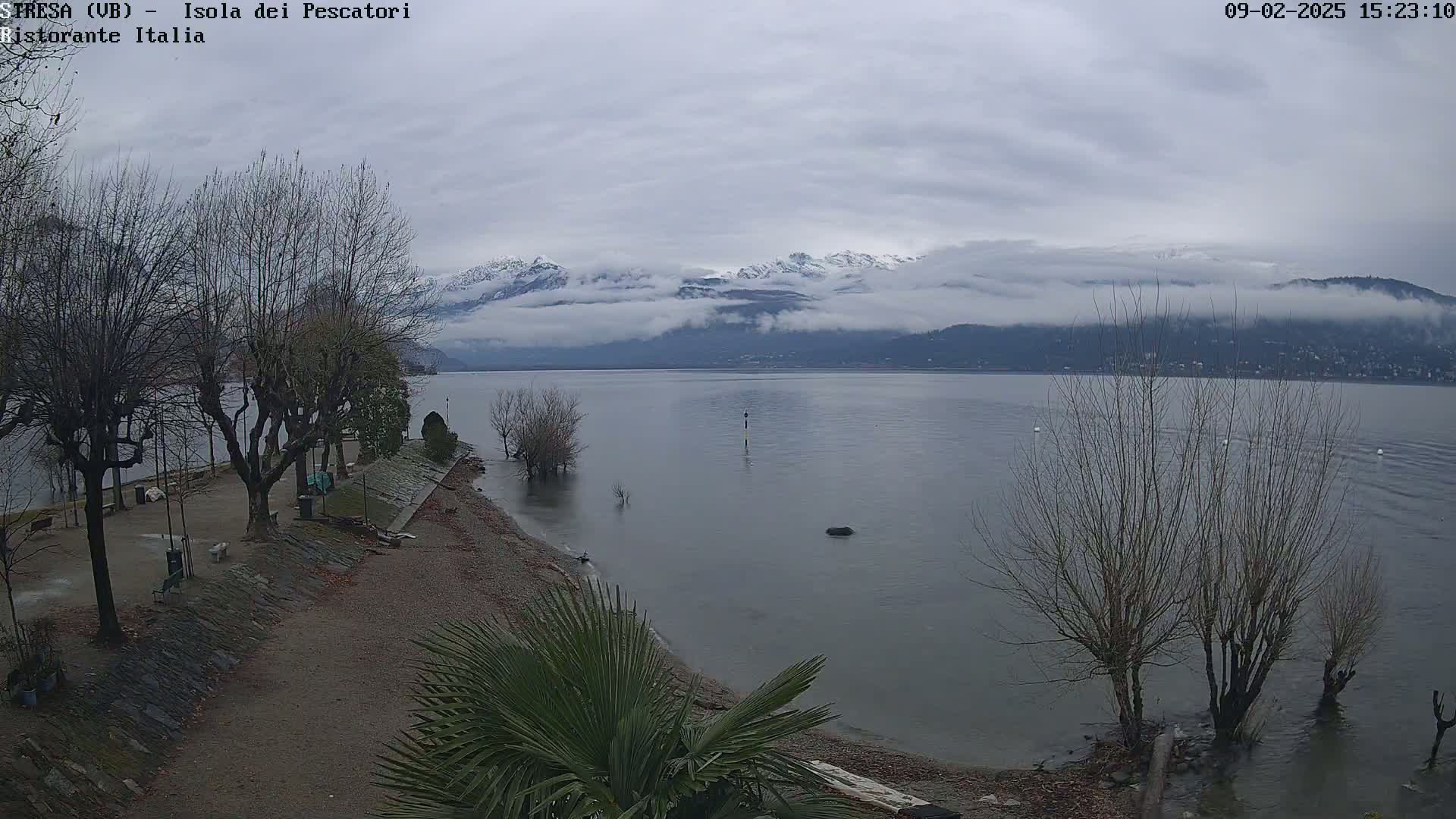 A calm lake with snow-capped mountains in the background under an overcast sky, viewed from a shoreline with bare trees and a small beach area.