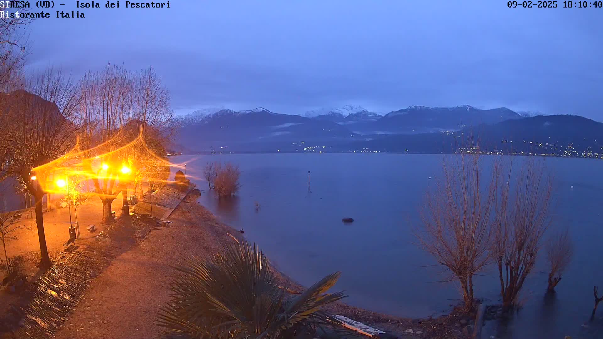 A twilight view shows a calm lake with snow-capped mountains in the distance, illuminated by streetlights along a shoreline path.