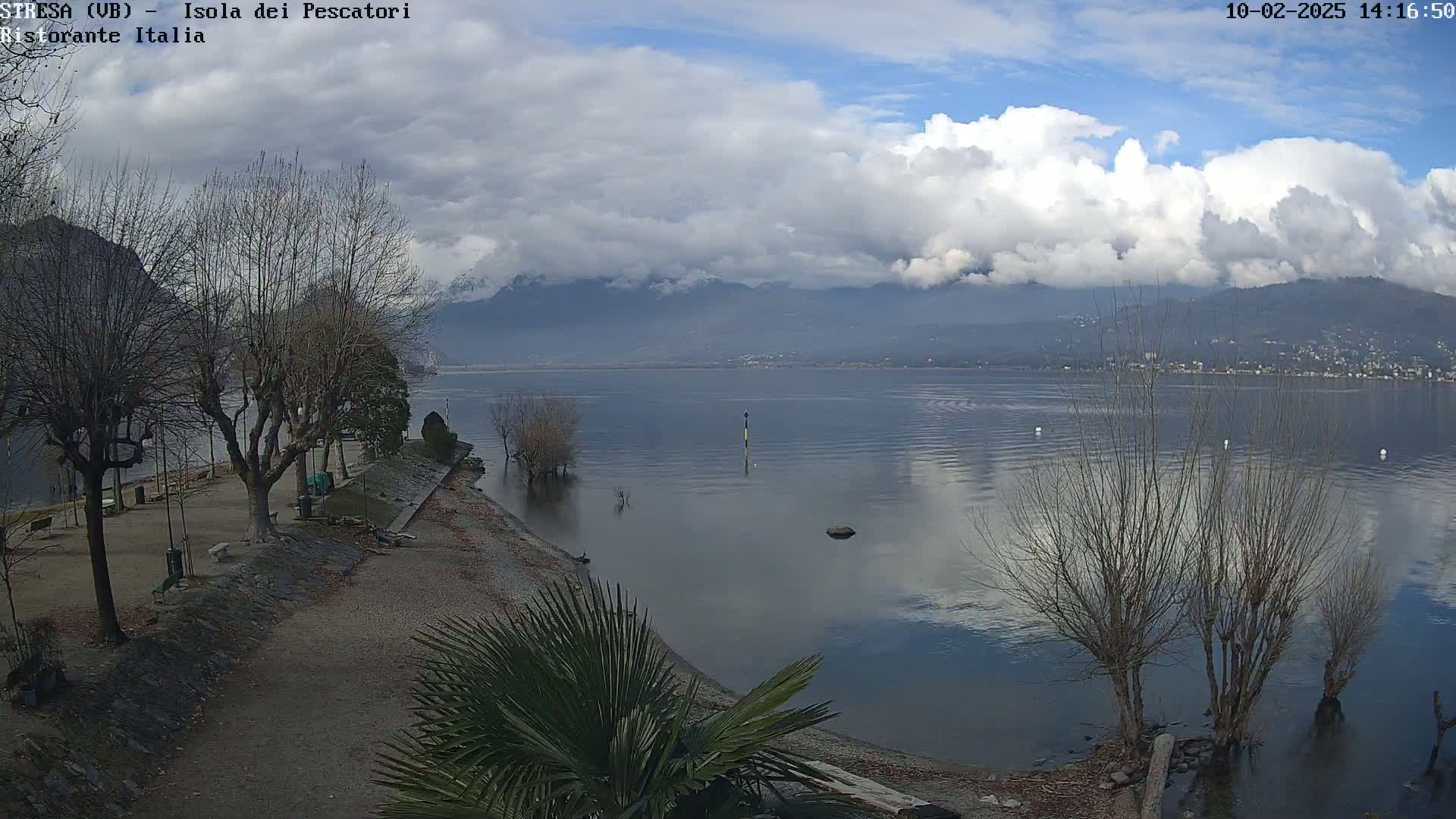 A partly cloudy day reveals a calm lake reflecting the sky and distant mountains, with bare trees lining a shoreline path.