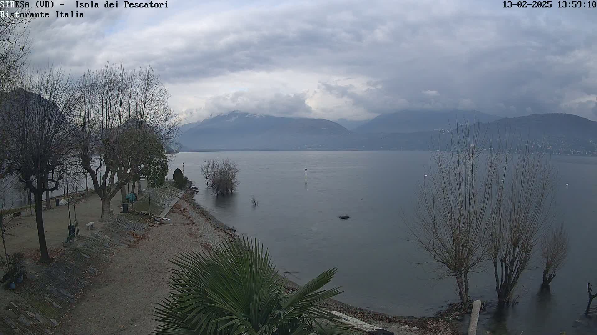 A calm lake is seen under a cloudy sky, with bare trees lining a shoreline path and mountains in the distance.