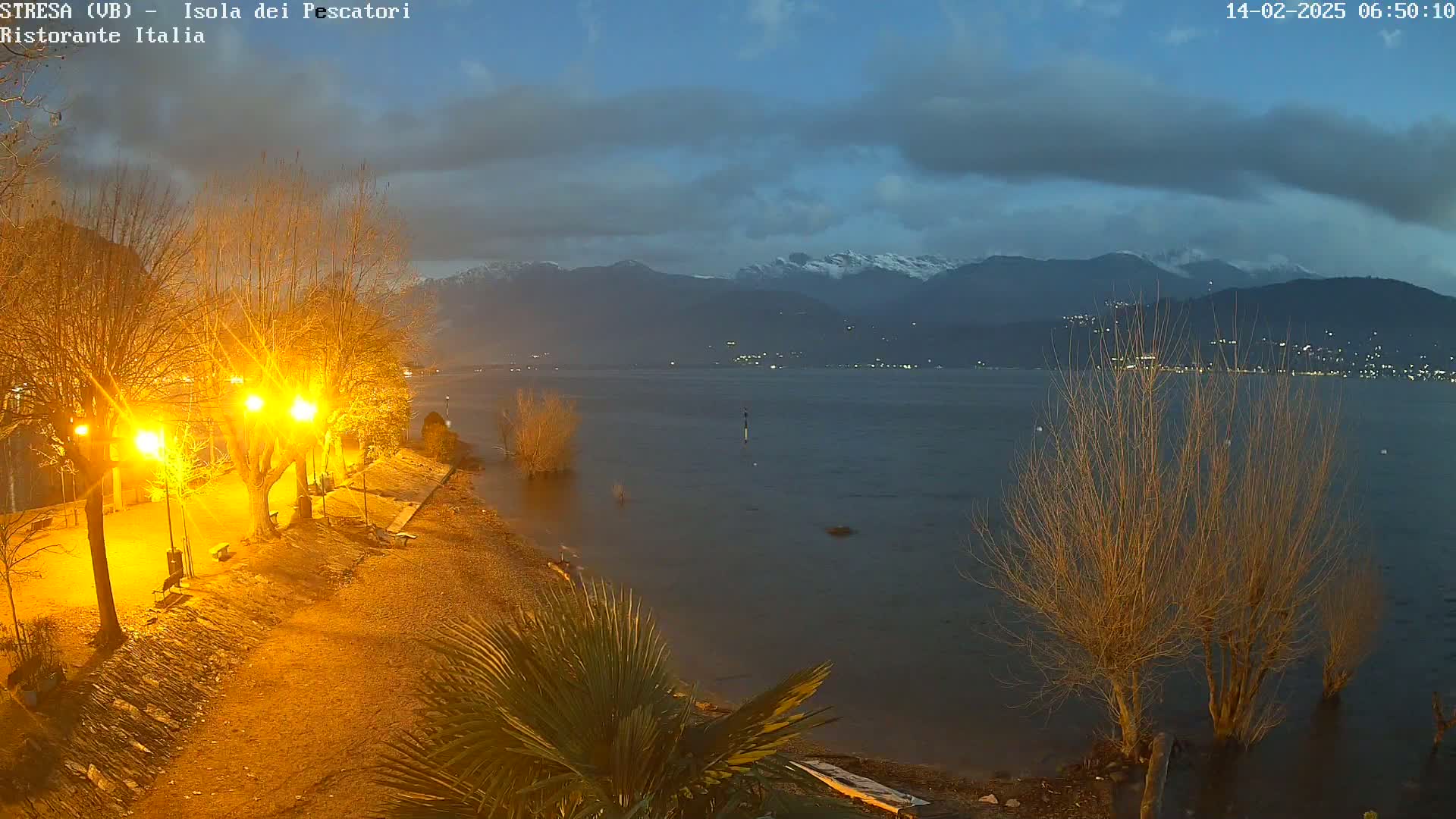 A nighttime lakeside view shows a paved path lit by streetlights, bare trees, and a calm lake with snow-capped mountains in the distance under a partly cloudy sky.