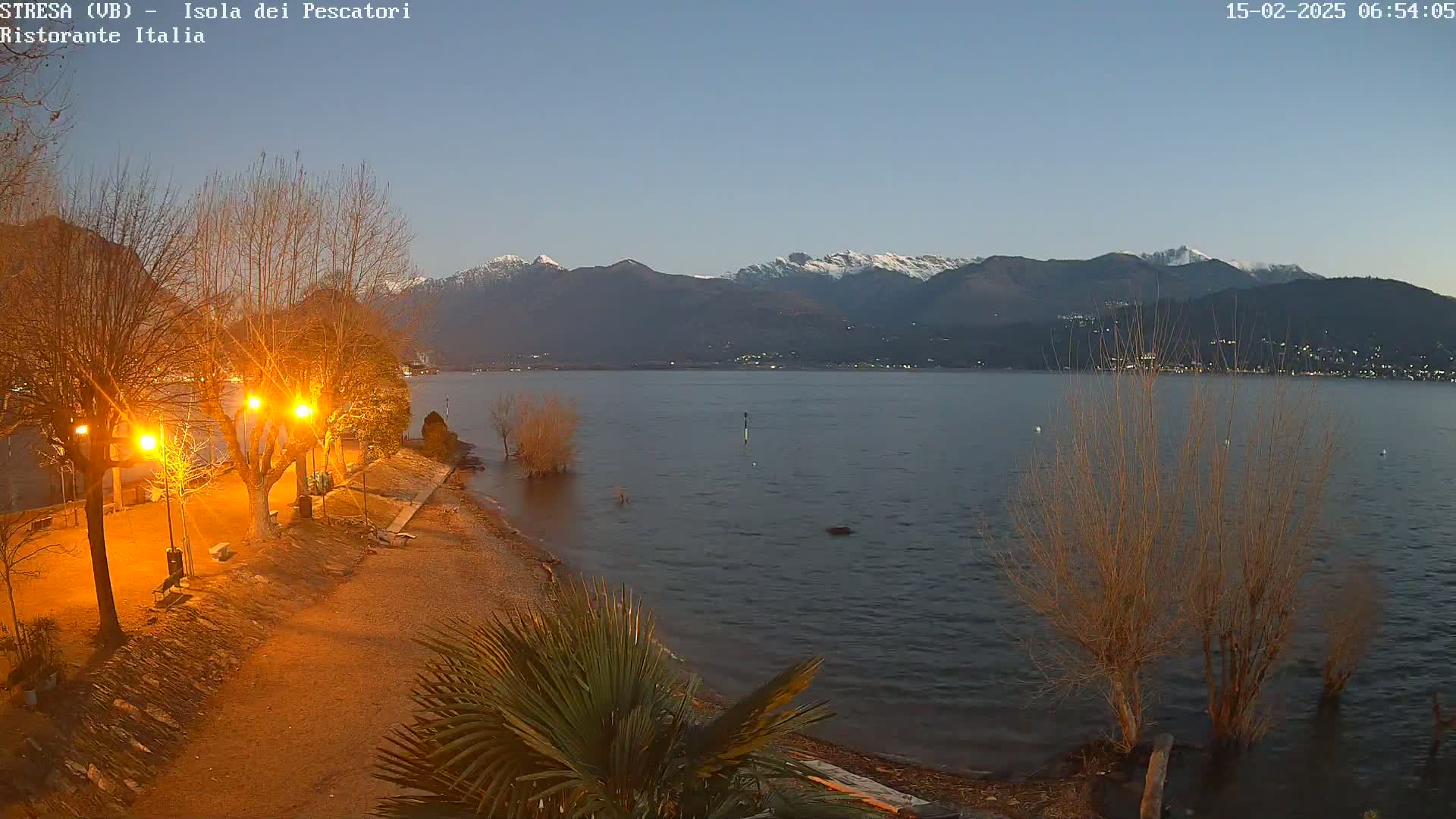 A dimly lit lakeshore scene at twilight shows bare trees illuminated by streetlights, a calm lake, and snow-capped mountains in the distance.