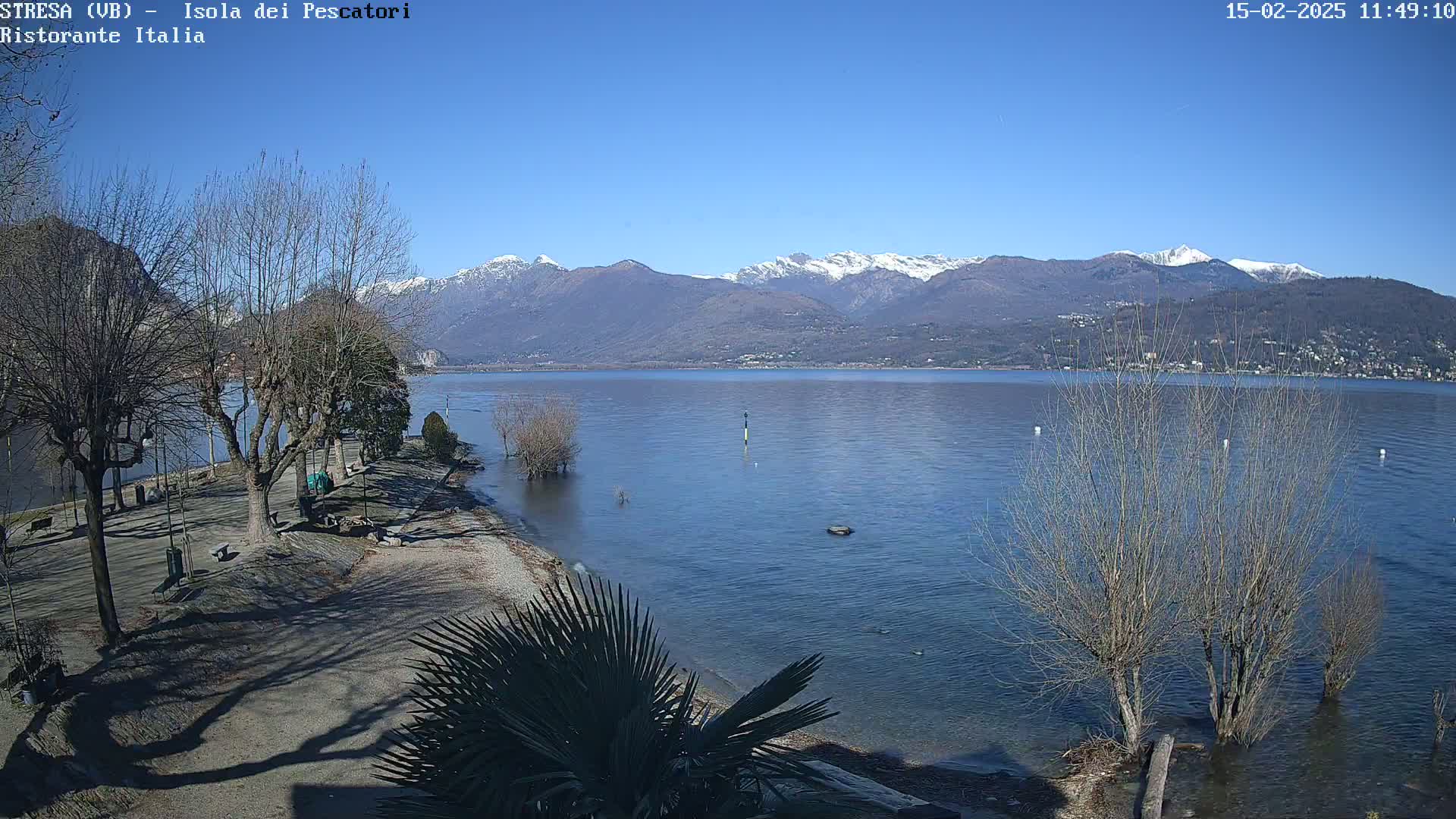 A tranquil lake scene under a clear blue sky shows snow-capped mountains in the distance, bare trees along the shoreline, and a calm body of water lapping at a pebble beach.