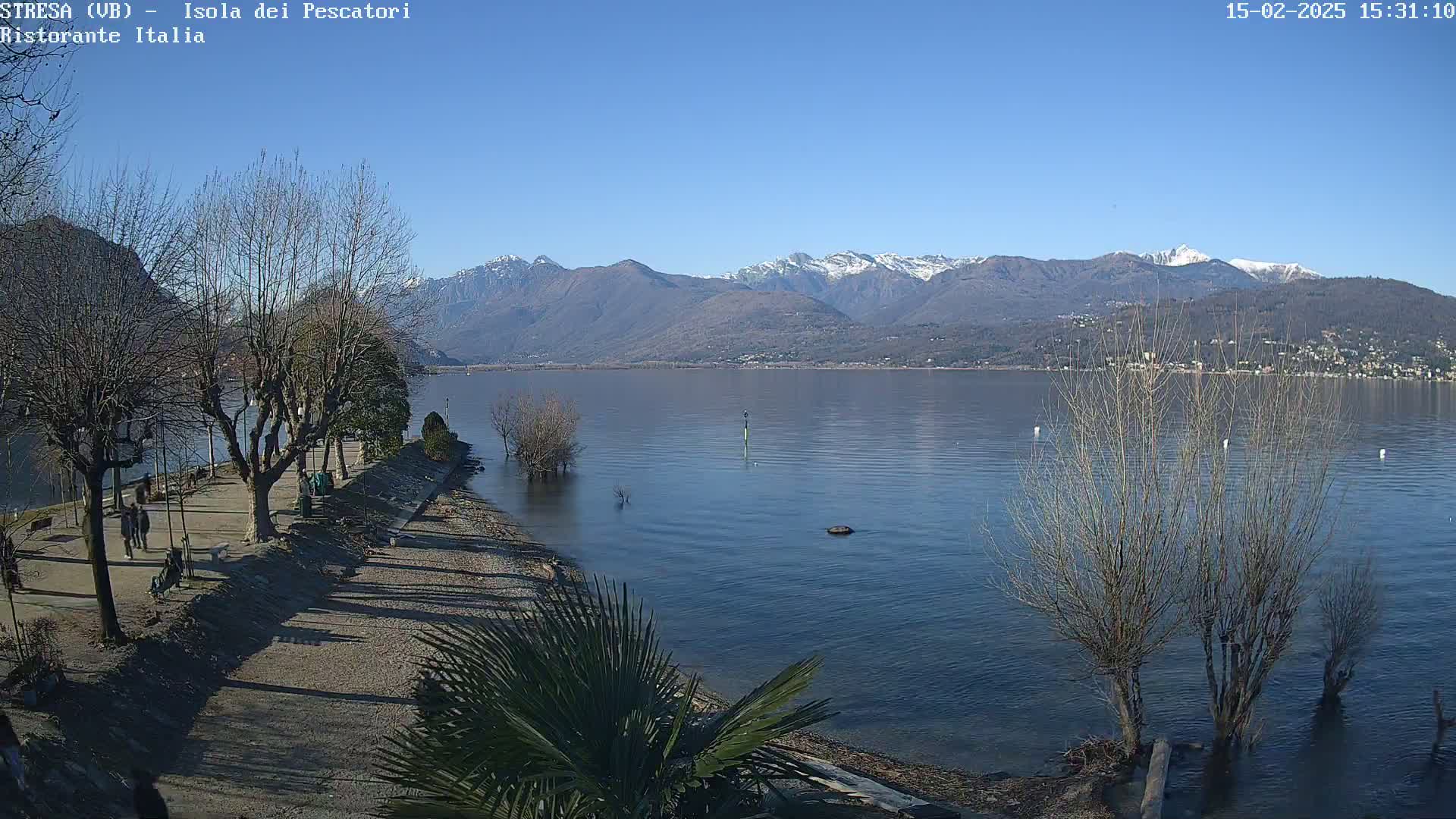 A calm lake with snow-capped mountains in the background on a sunny day, seen from a lakeside promenade with bare trees and a few people walking.