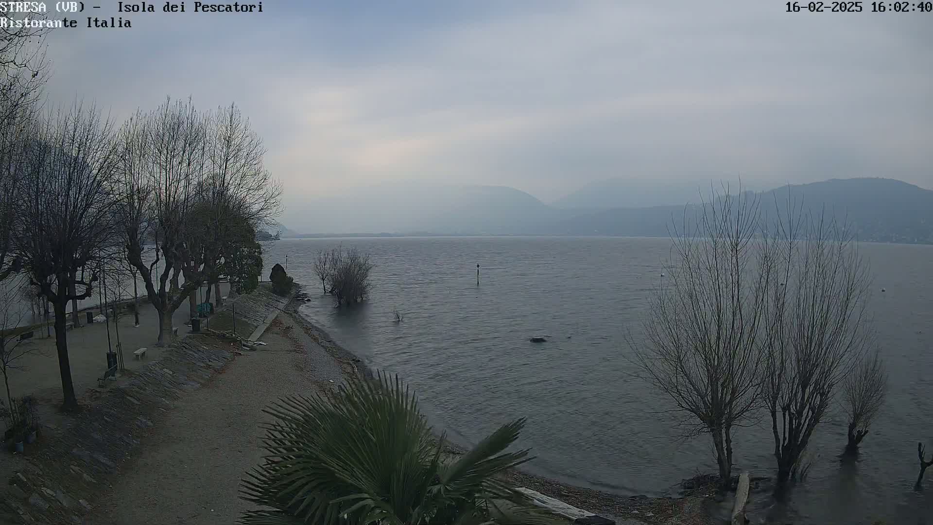 A mostly gray, overcast day shows a calm lake with bare trees lining a pebble shore, mountains in the background.