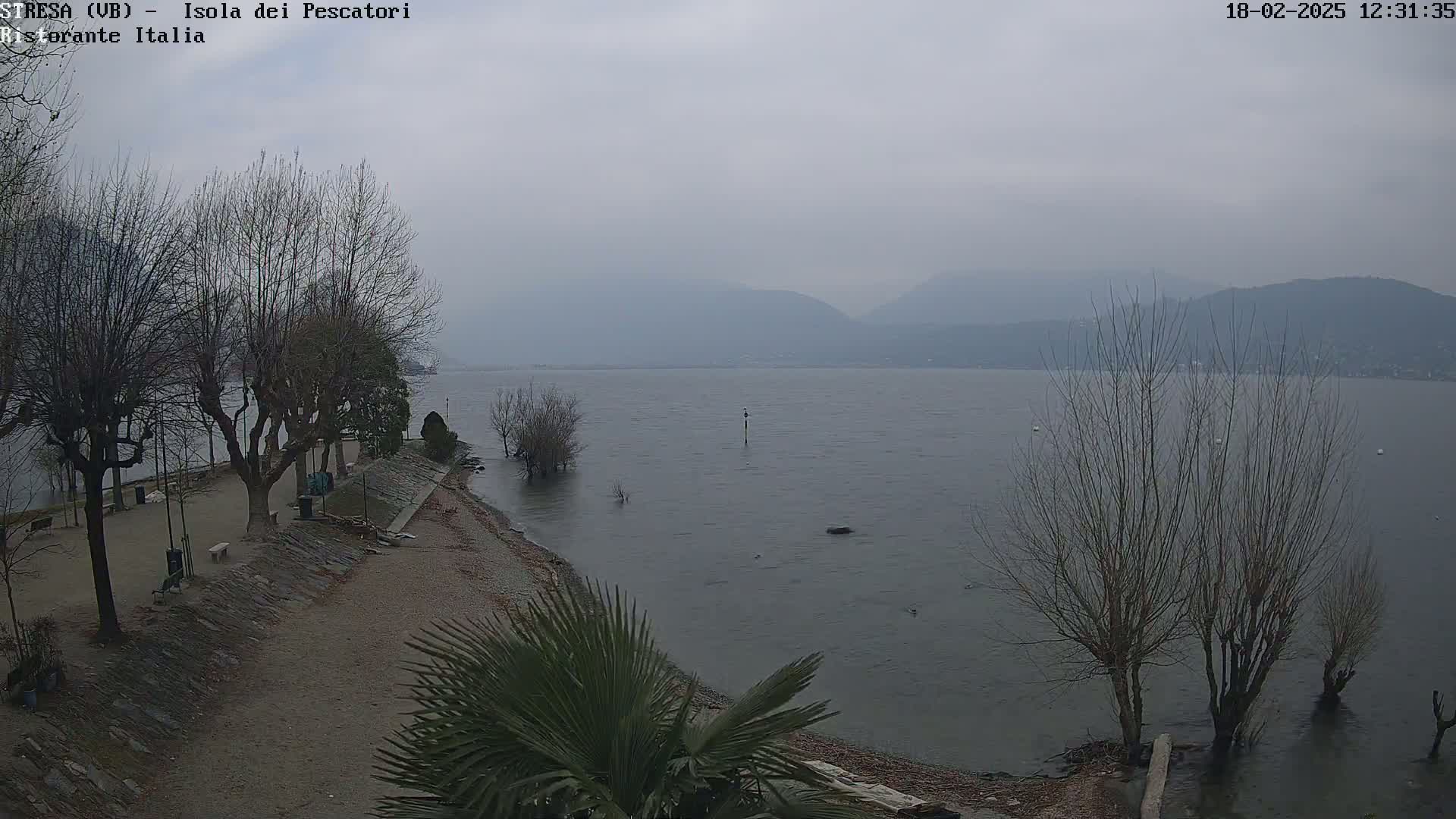 A gray, overcast day shows a calm lake with bare trees lining a shoreline, where a sandy beach meets a stone retaining wall and a paved area with some benches.